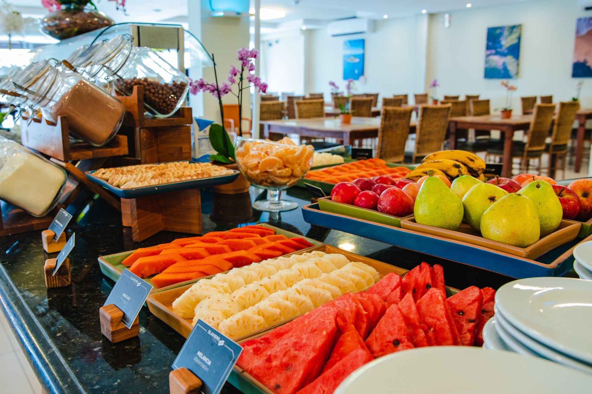 Mesa de buffet com bandejas de frutas variadas, incluindo melancia, melão, laranjas e peras, em uma sala de jantar iluminada.