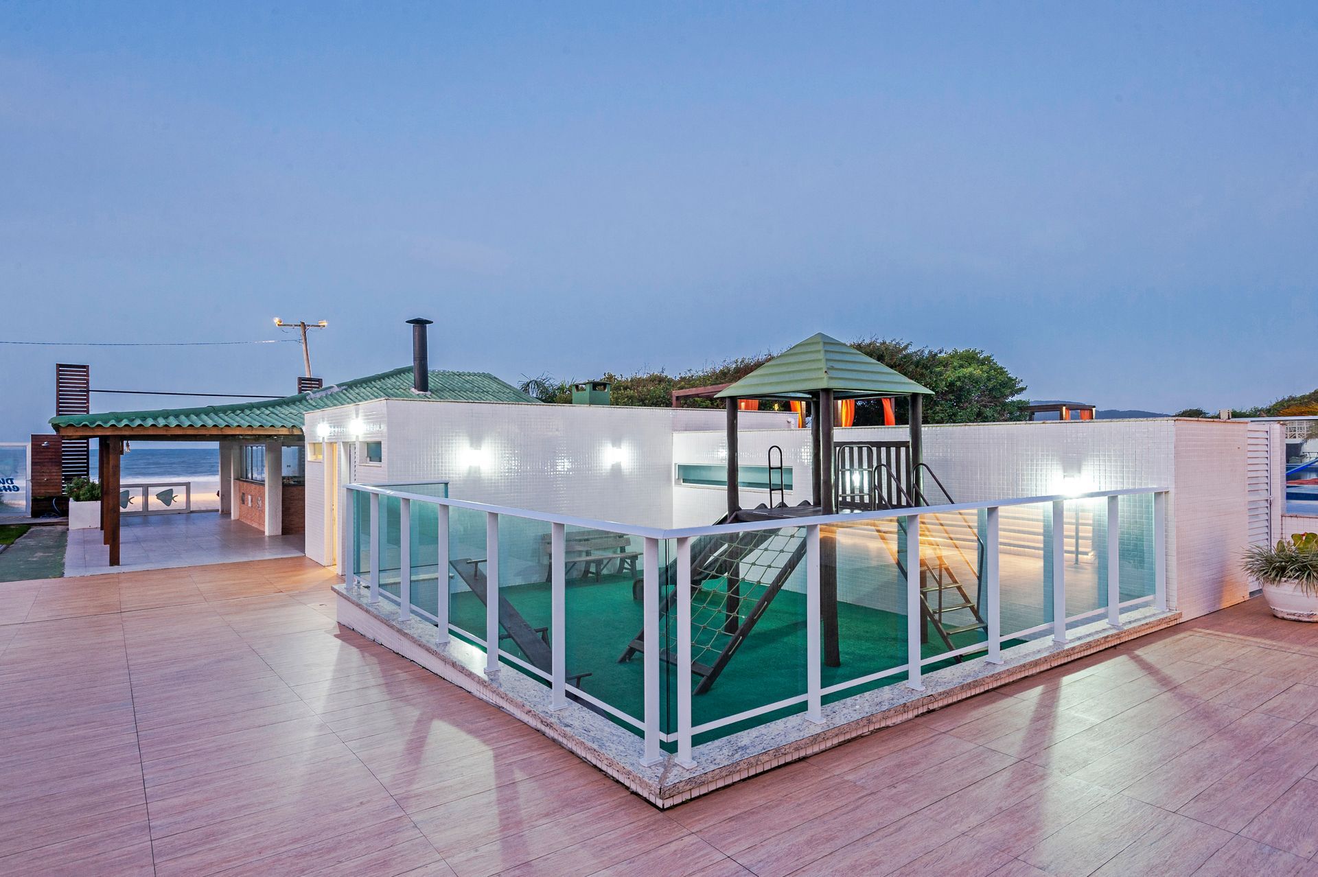 Pavilhão moderno à beira-mar com paredes de vidro e telhado verde ao entardecer, ao lado de uma passarela de azulejos molhada.