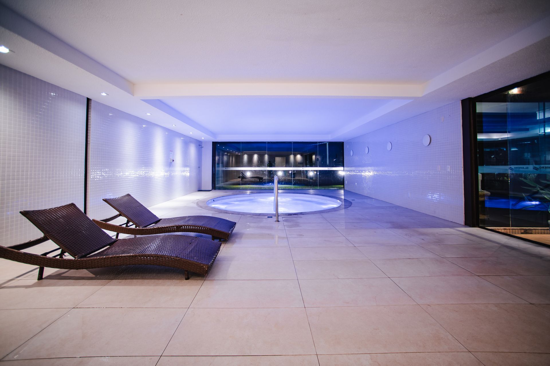 Piscina coberta na cobertura com espreguiçadeiras, iluminação azul e vista da cidade através de paredes de vidro.