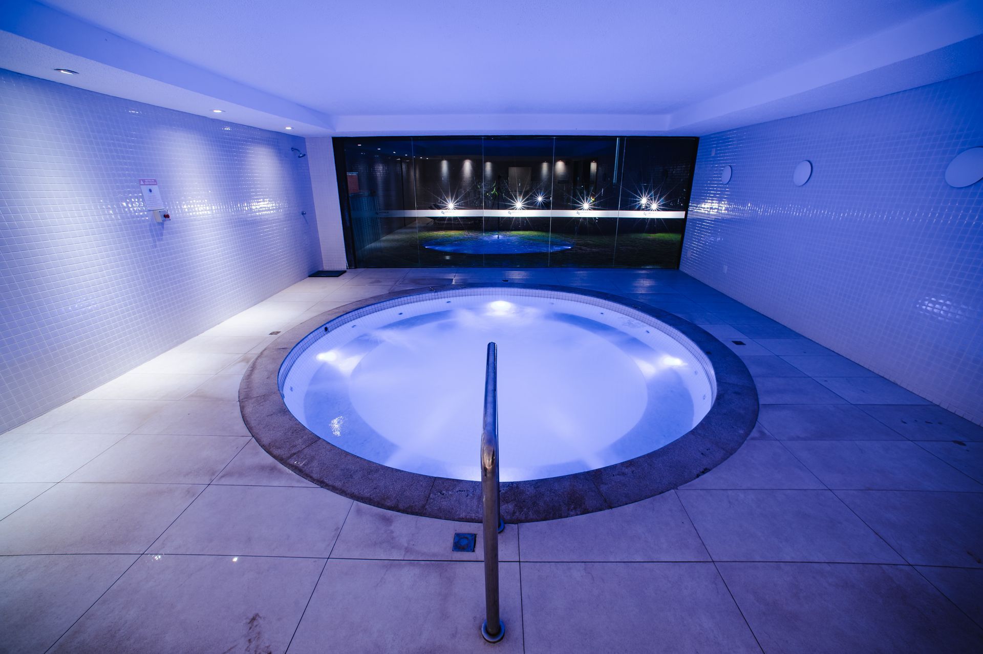 Banheira de hidromassagem interna com iluminação azul, paredes revestidas de azulejos e corrimão de metal.