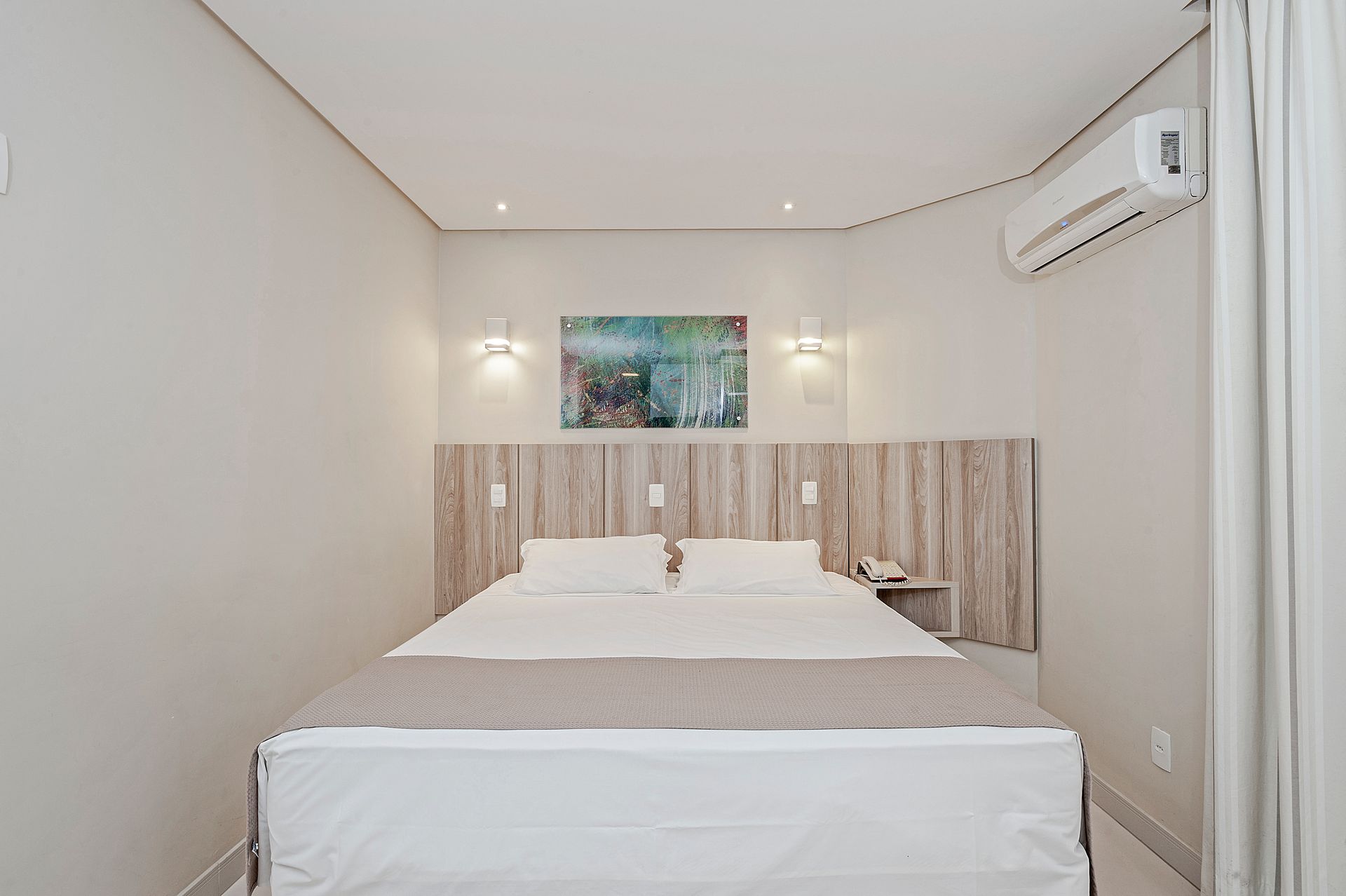 Quarto minimalista e organizado com cama de casal, cabeceira de madeira, obras de arte na parede e ar condicionado.