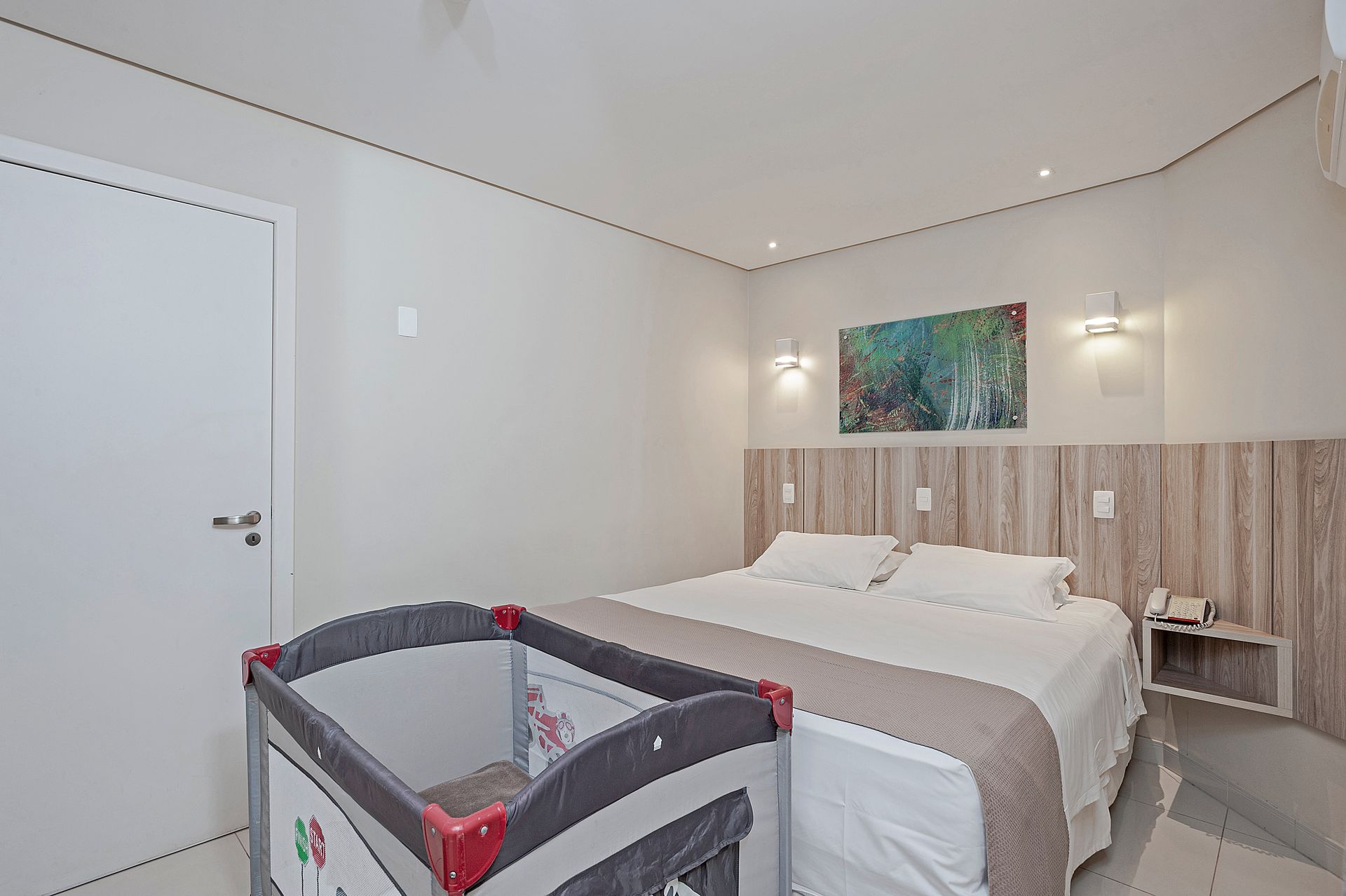 Quarto iluminado com cama, obras de arte na parede, luminárias de cabeceira e um berço em primeiro plano.