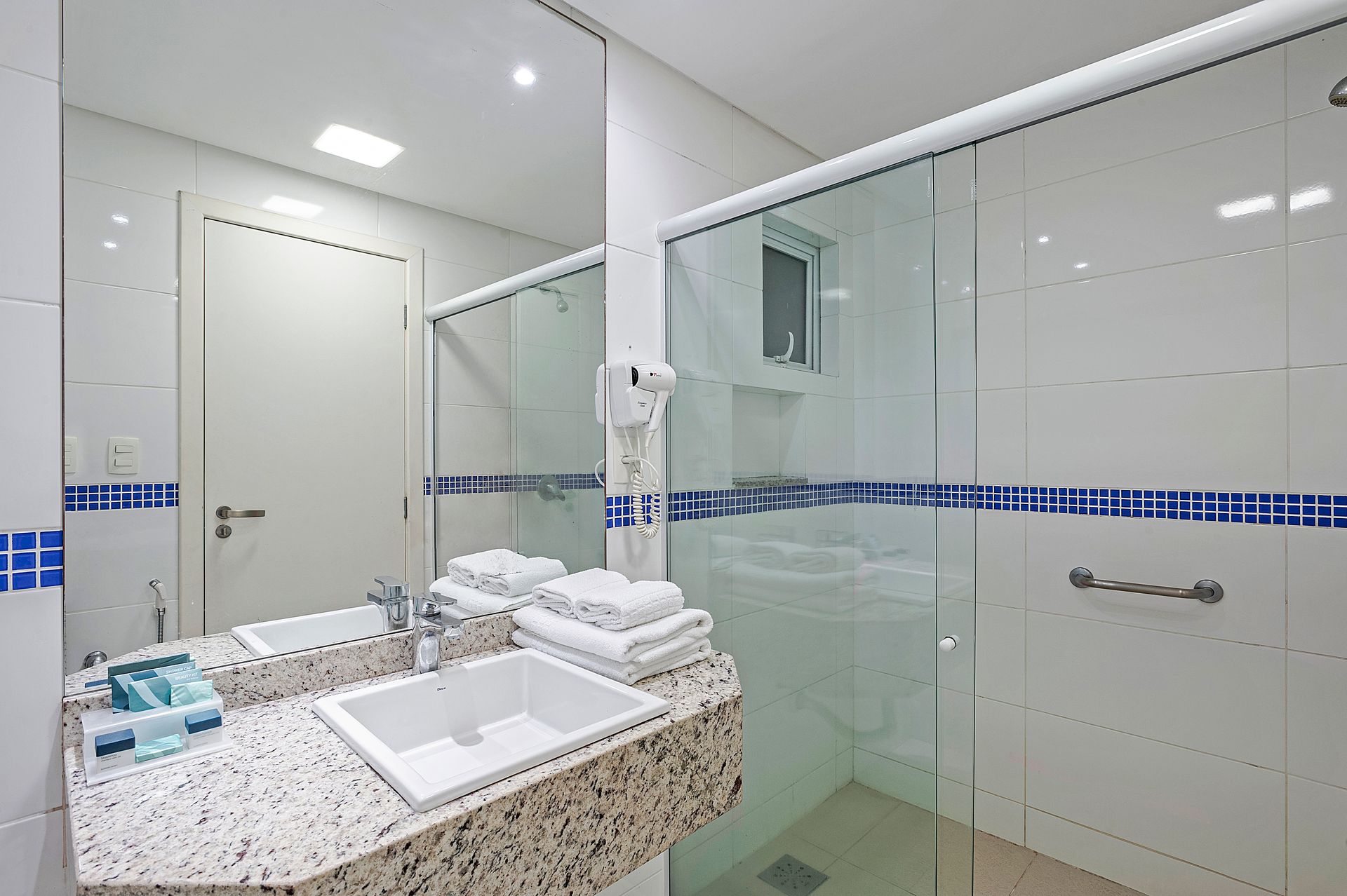 Banheiro moderno com box de vidro, pia branca e faixa decorativa de azulejos azuis.