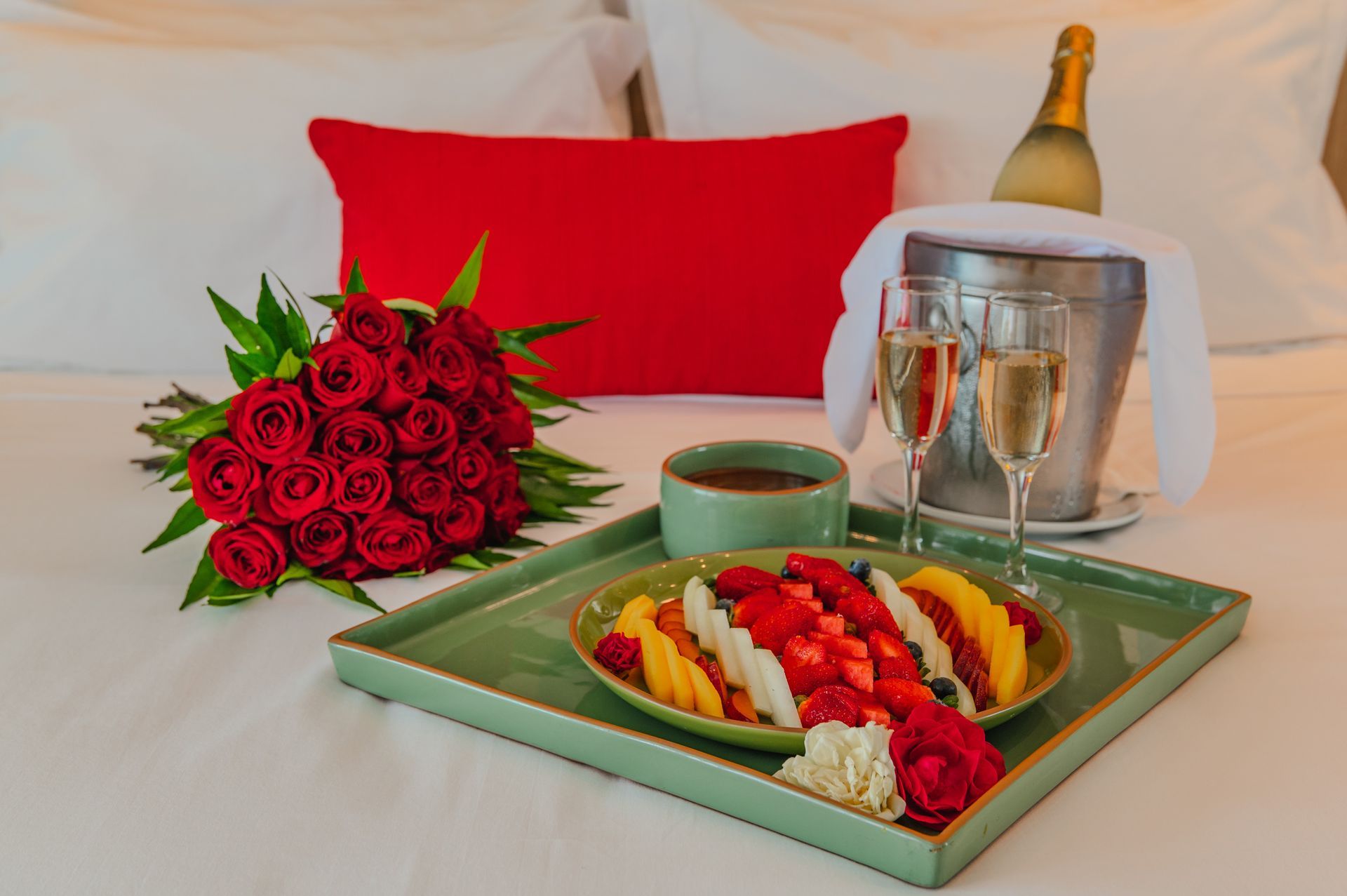 Bandeja de café da manhã romântica com frutas, champanhe, rosas e uma almofada vermelha sobre uma cama branca.