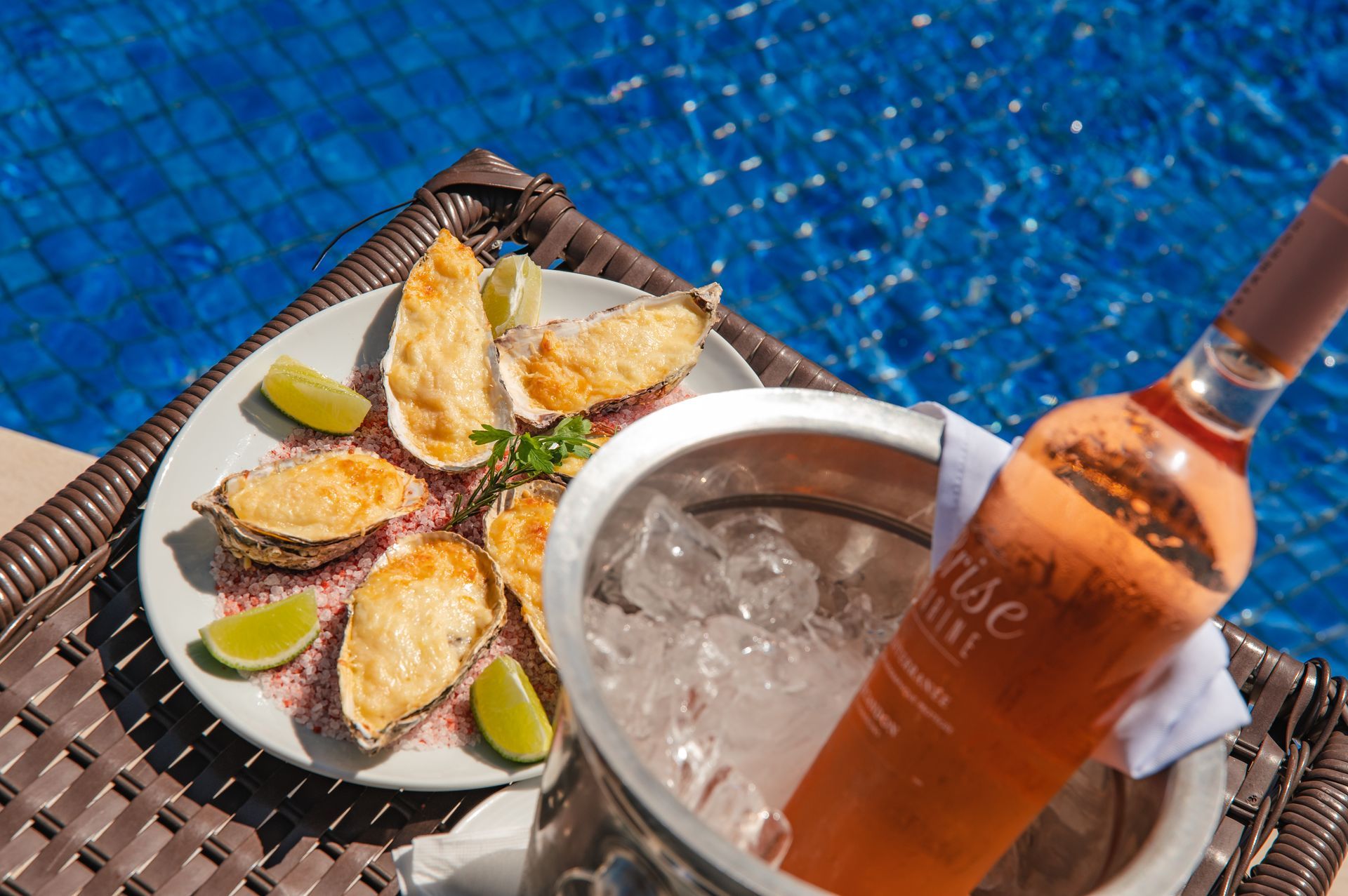 Mesa à beira da piscina com bebida gelada, garrafa e aperitivos servidos no prato, junto à água azul.