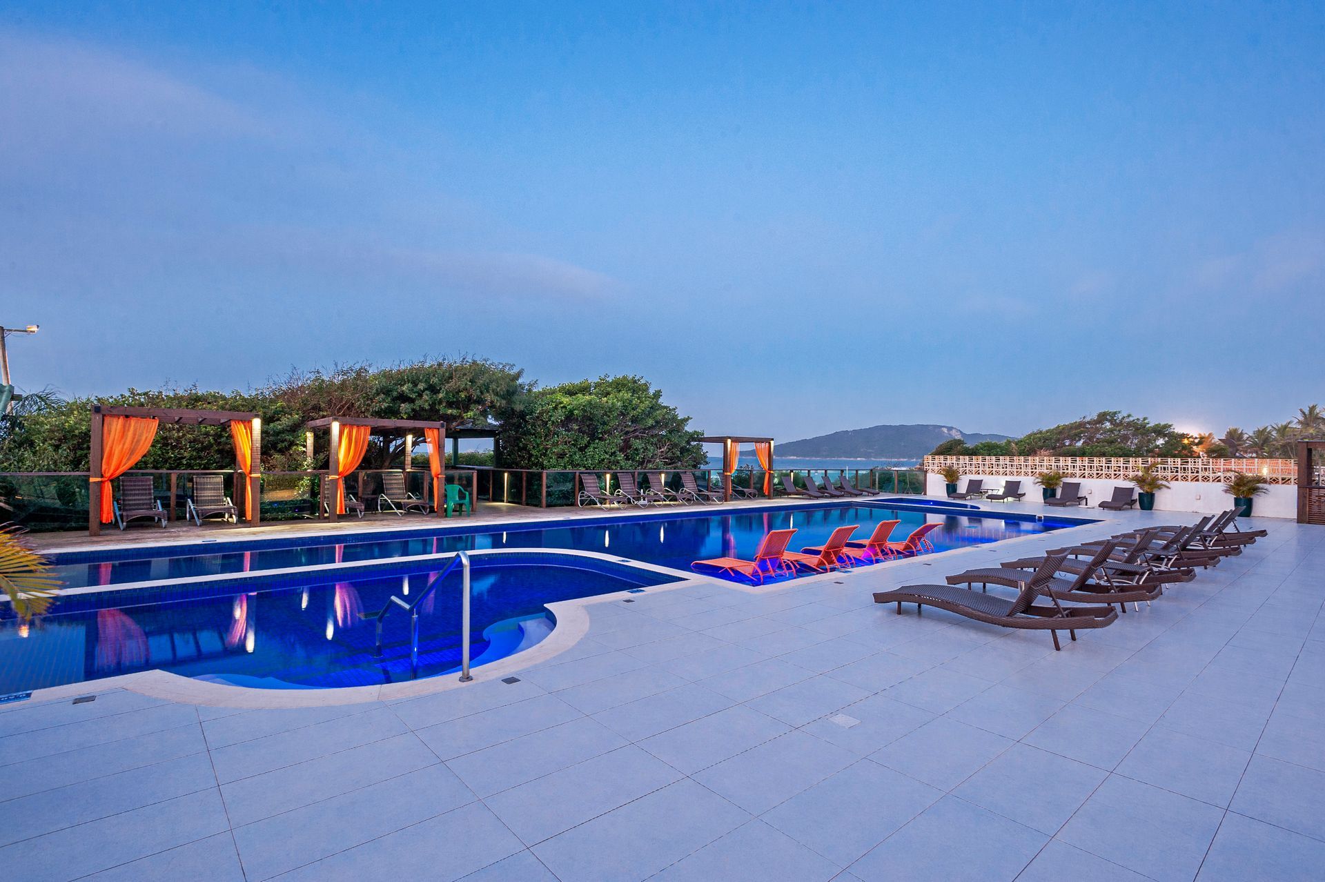 Piscina externa do resort ao entardecer, com espreguiçadeiras, cabanas e água azul sob um céu em tons pastel.