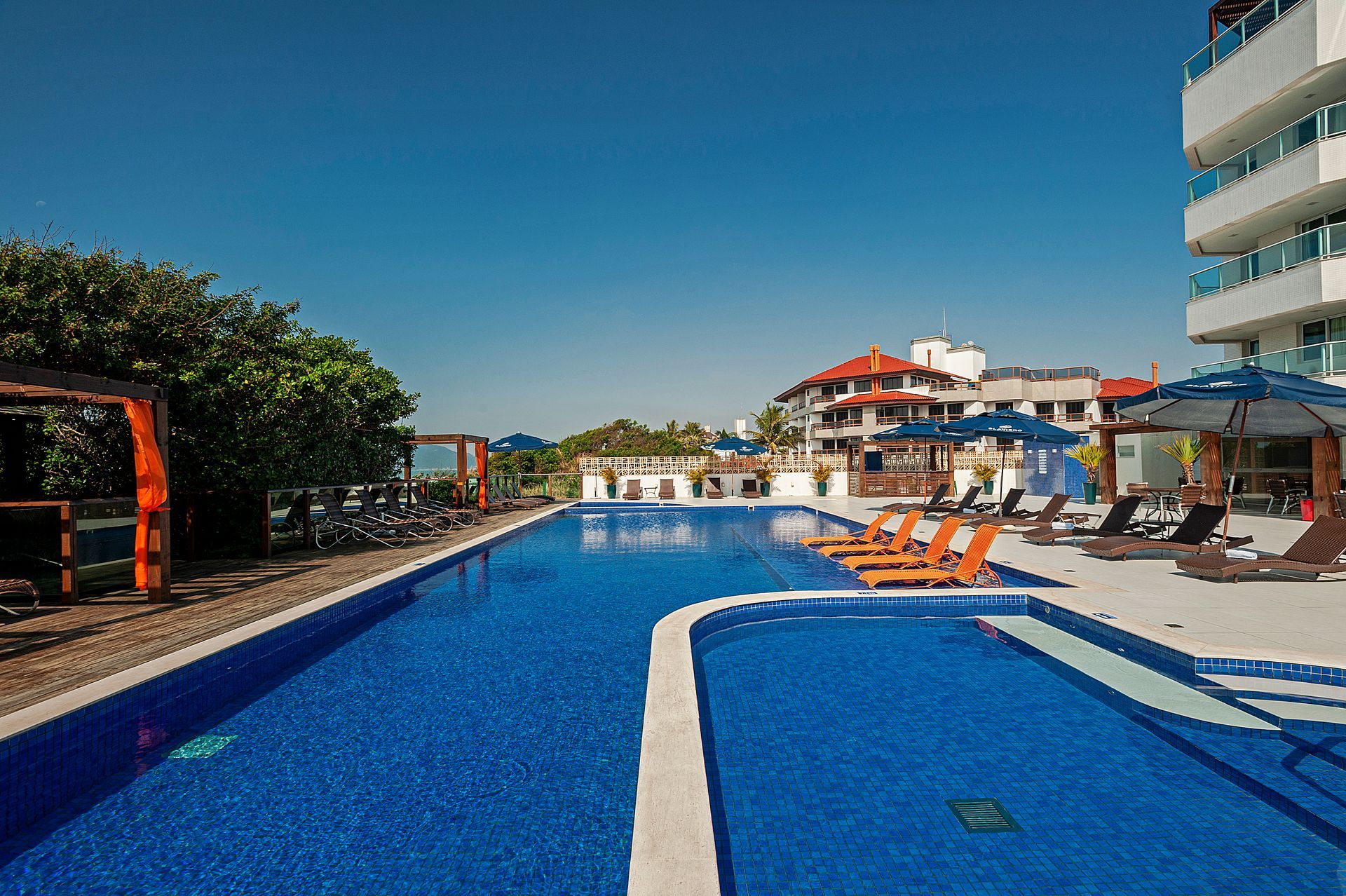 Piscina de resort com água azul, espreguiçadeiras laranja e guarda-sóis sob um céu claro.