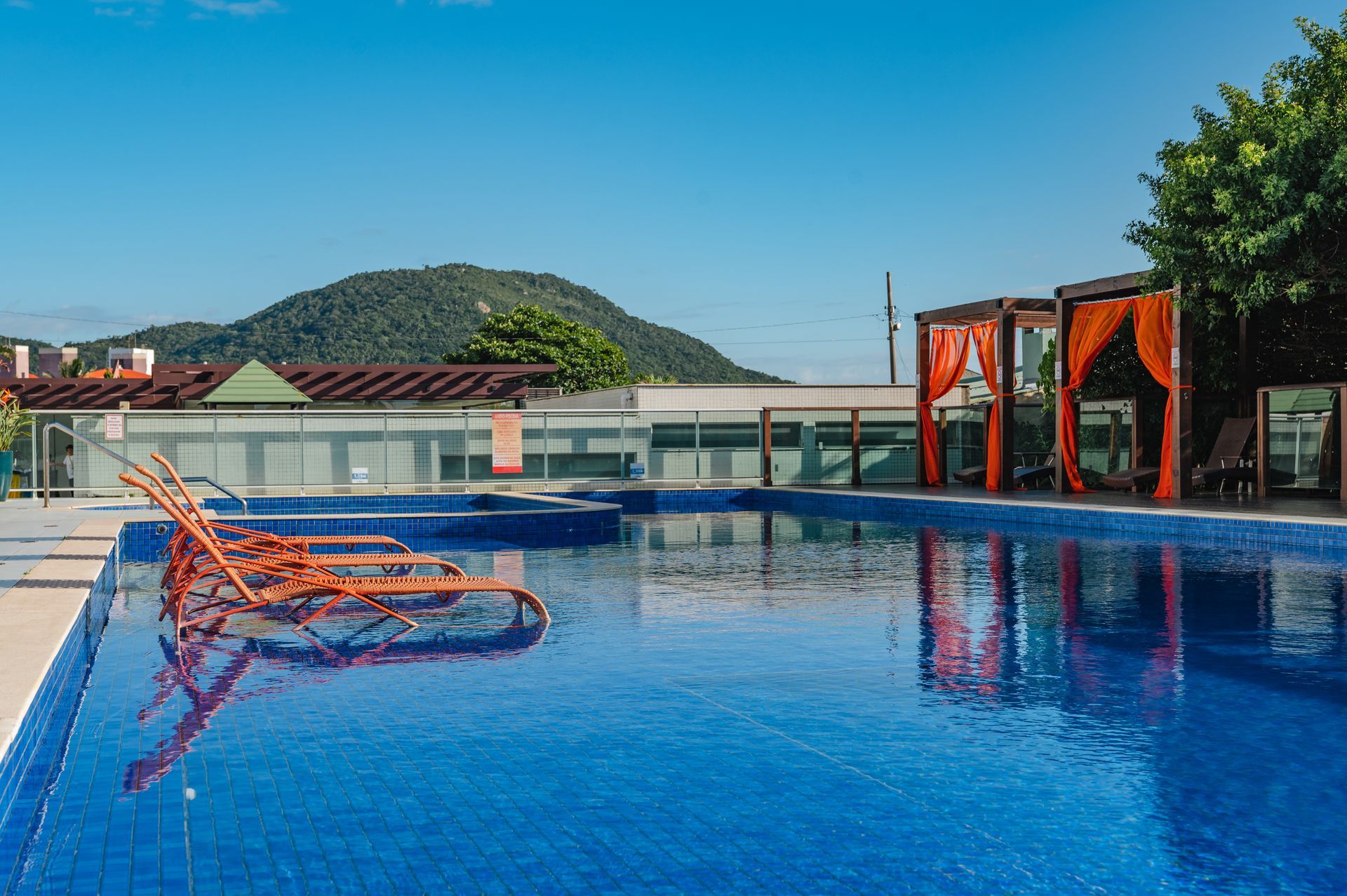 Piscina infinita com água azul, espreguiçadeiras laranja e cabanas sombreadas com vista para o oceano e as colinas.