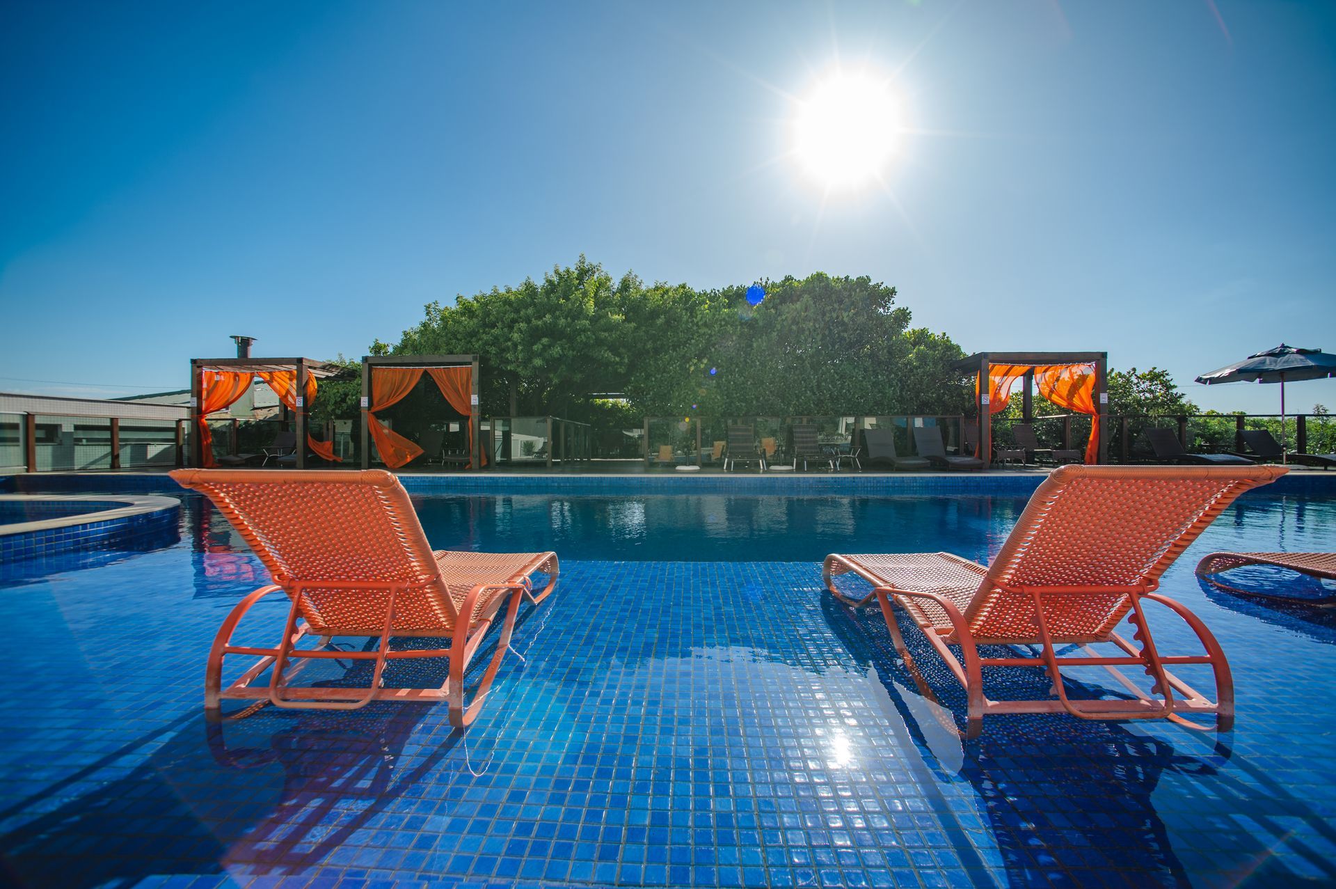 Espreguiçadeiras laranja à beira de uma piscina azul sob um sol radiante, com árvores e cabanas ao fundo.