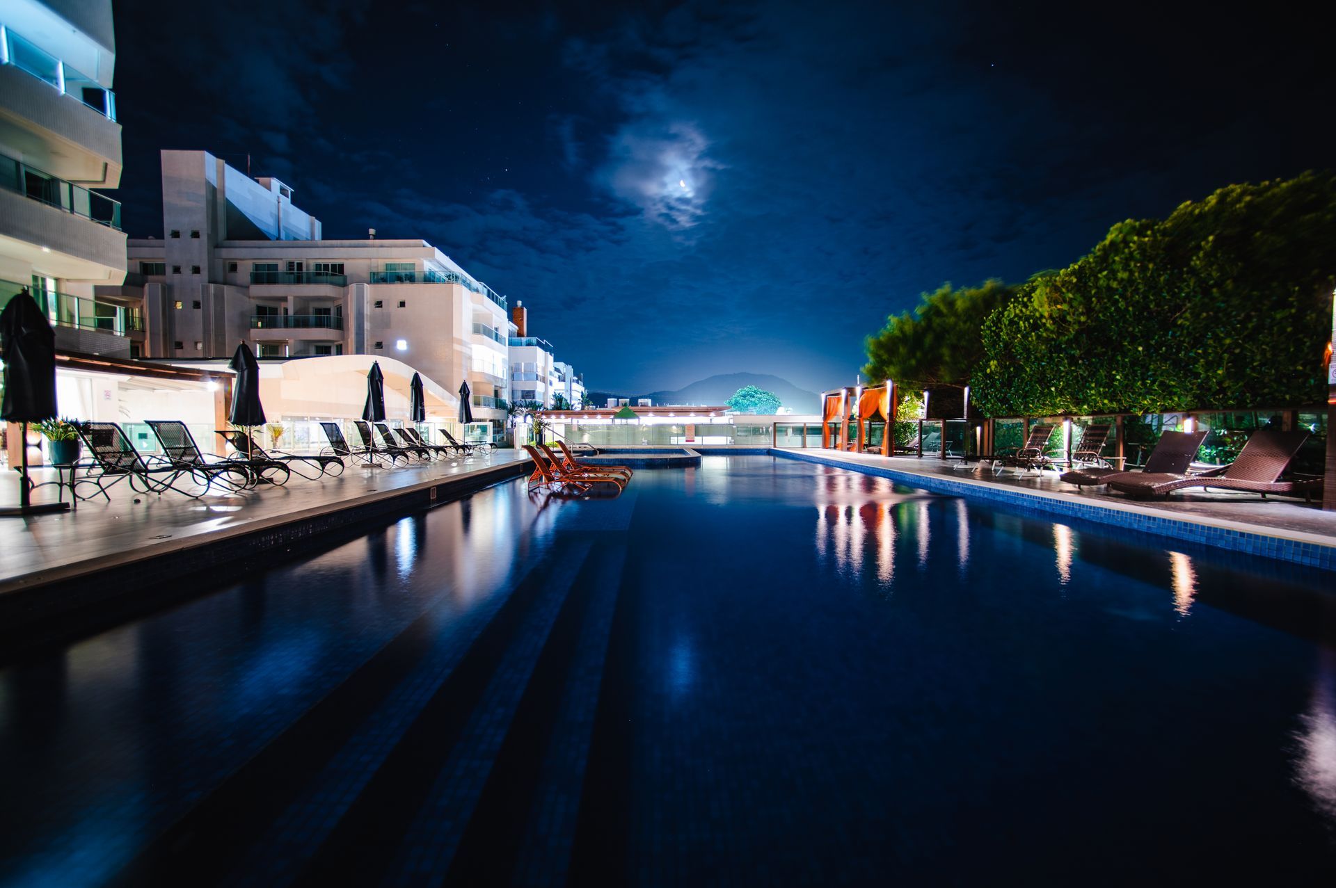 Paisagem urbana noturna à beira-mar com edifícios iluminados, calçadão arborizado e reflexos na água calma.