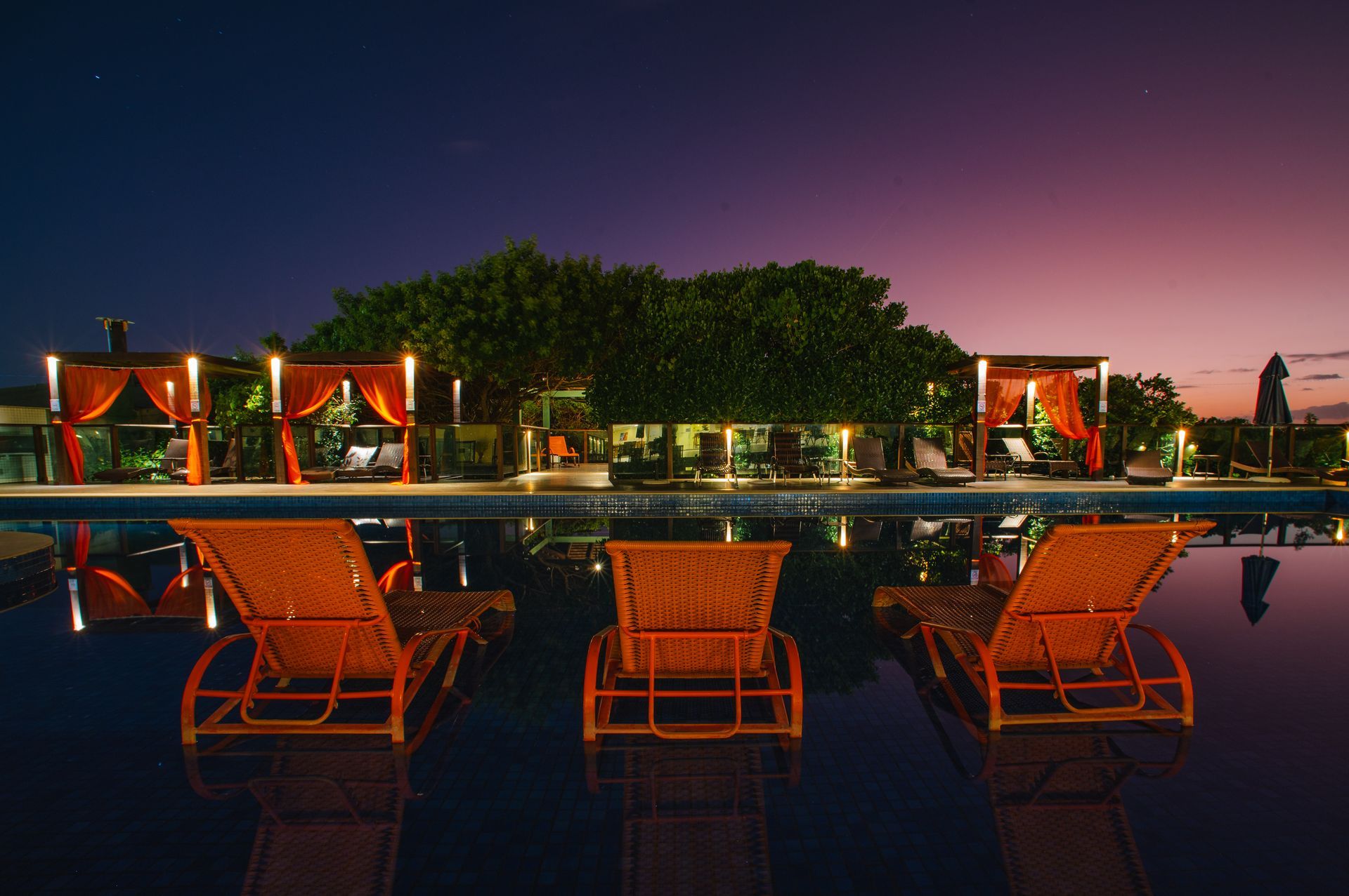 Espreguiçadeiras à beira da piscina ao entardecer, com cabanas iluminadas e um céu rosa-azulado ao pôr do sol.