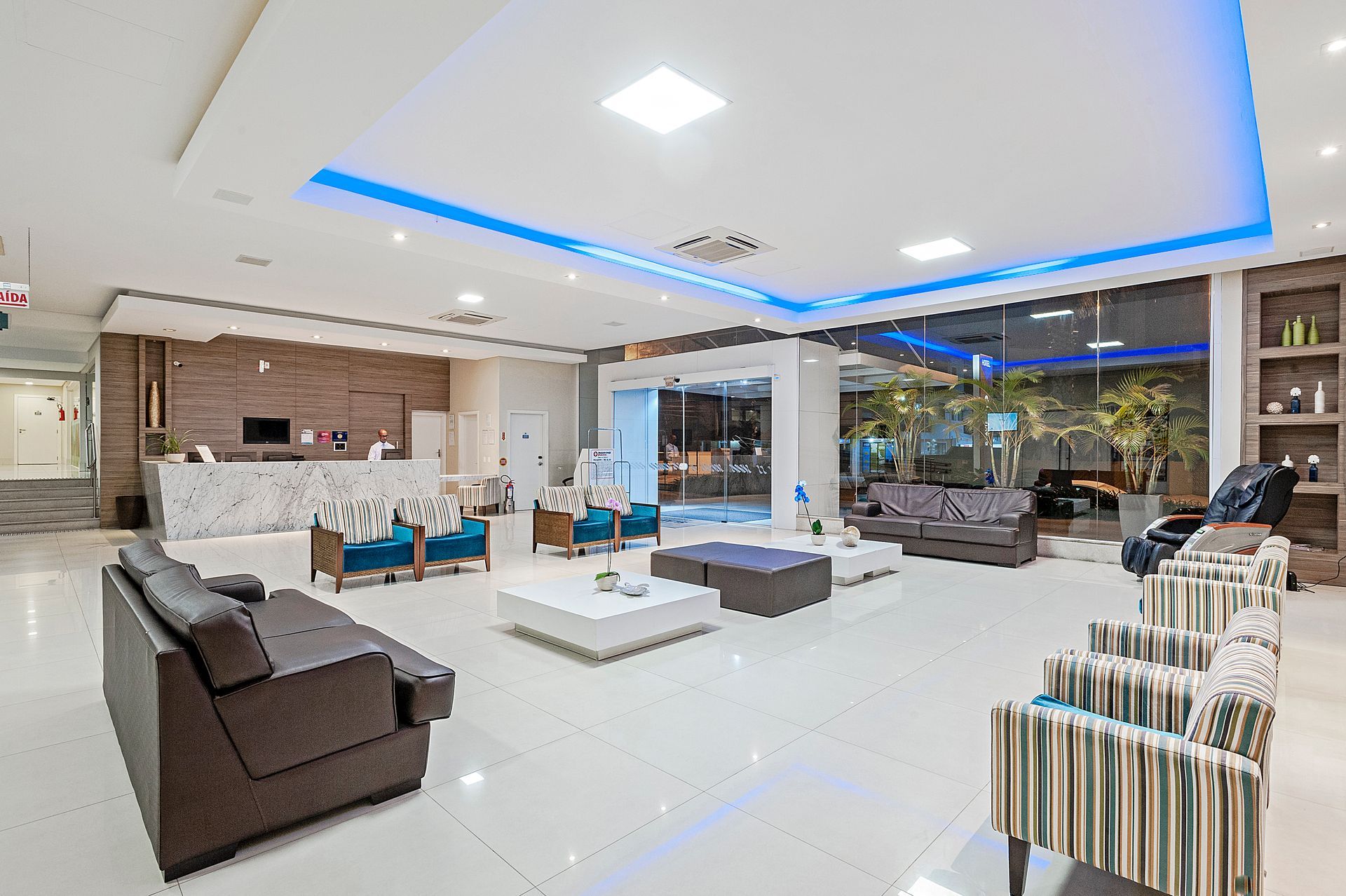 Sala de estar moderna com piso de azulejo branco, sofás marrons, luminárias de LED azuis no teto e janelas amplas.