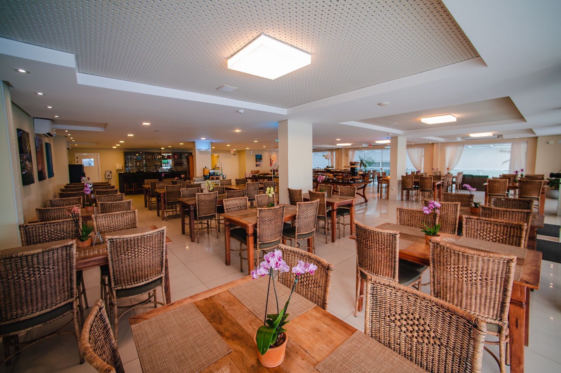 Sala de jantar de restaurante iluminada com cadeiras de vime, mesas de madeira e iluminação aconchegante no teto.