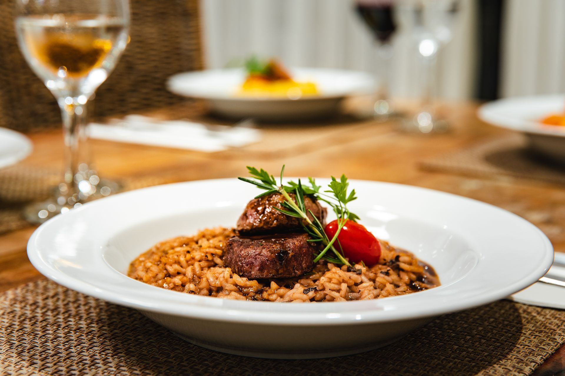Bife servido com arroz e guarnição de tomate em uma mesa de jantar, com uma taça de vinho ao fundo.
