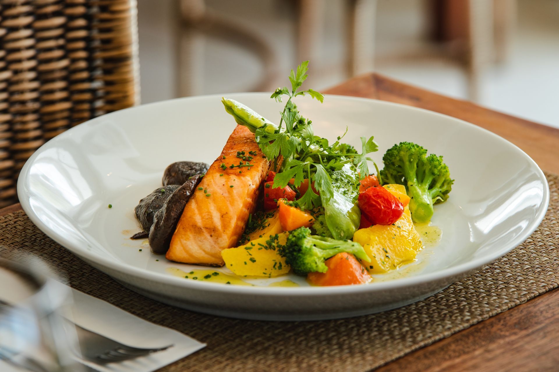 Filé de salmão com legumes e verduras servido em um prato branco sobre uma mesa de madeira.