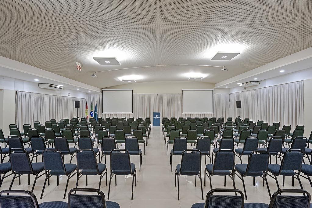Sala de conferências vazia com fileiras de cadeiras voltadas para uma tela de projeção e um pódio.