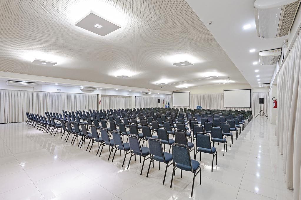Sala de conferências vazia com fileiras de cadeiras azuis e uma grande tela de projeção.