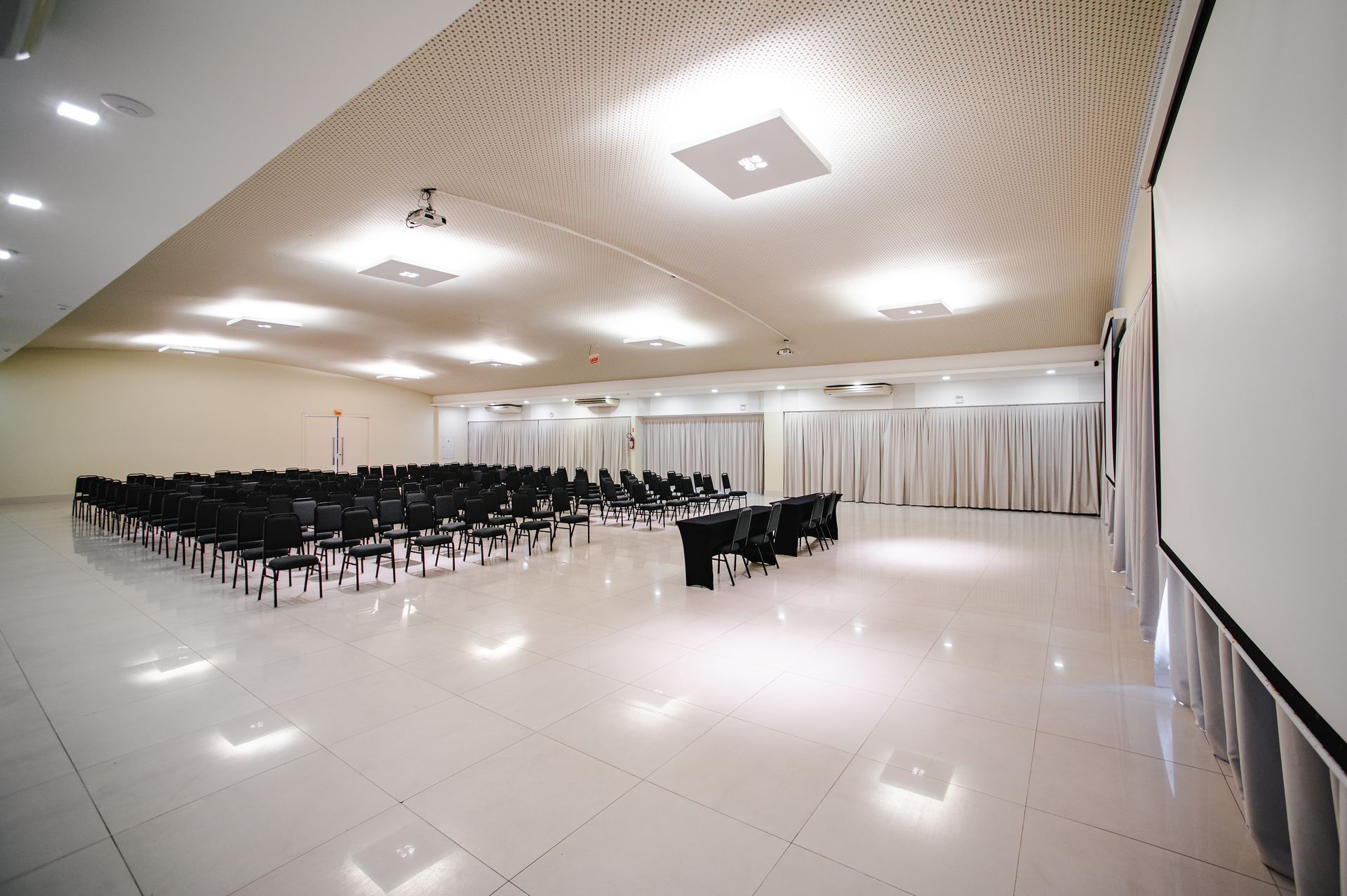 Sala de conferências vazia com fileiras de cadeiras pretas, piso branco brilhante, teto ornamentado e uma tela de projeção.