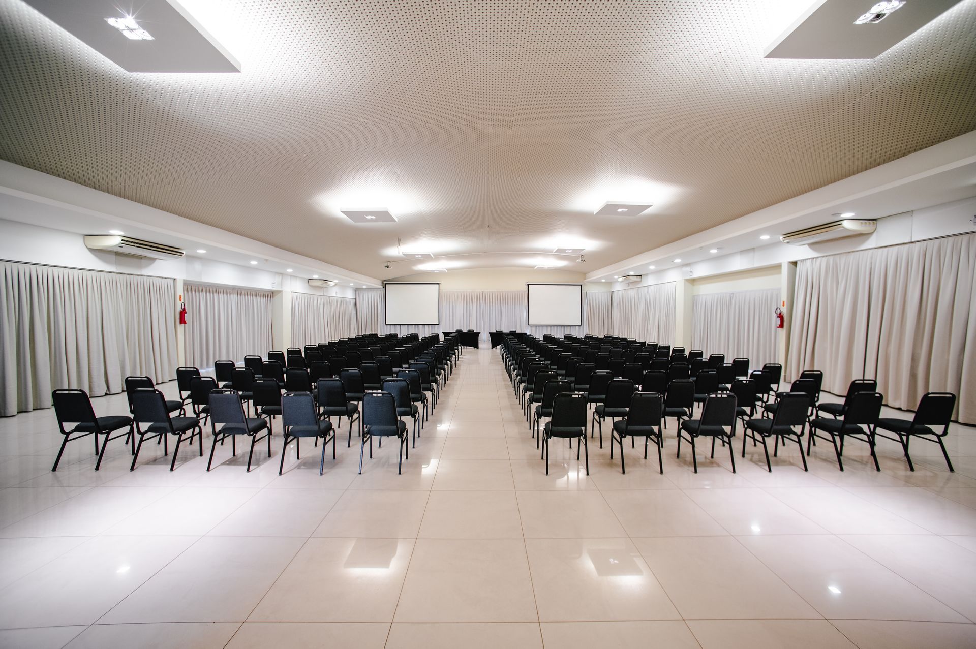 Salão de banquetes vazio com fileiras de cadeiras pretas e paredes cobertas por cortinas brancas sob luzes brilhantes no teto.