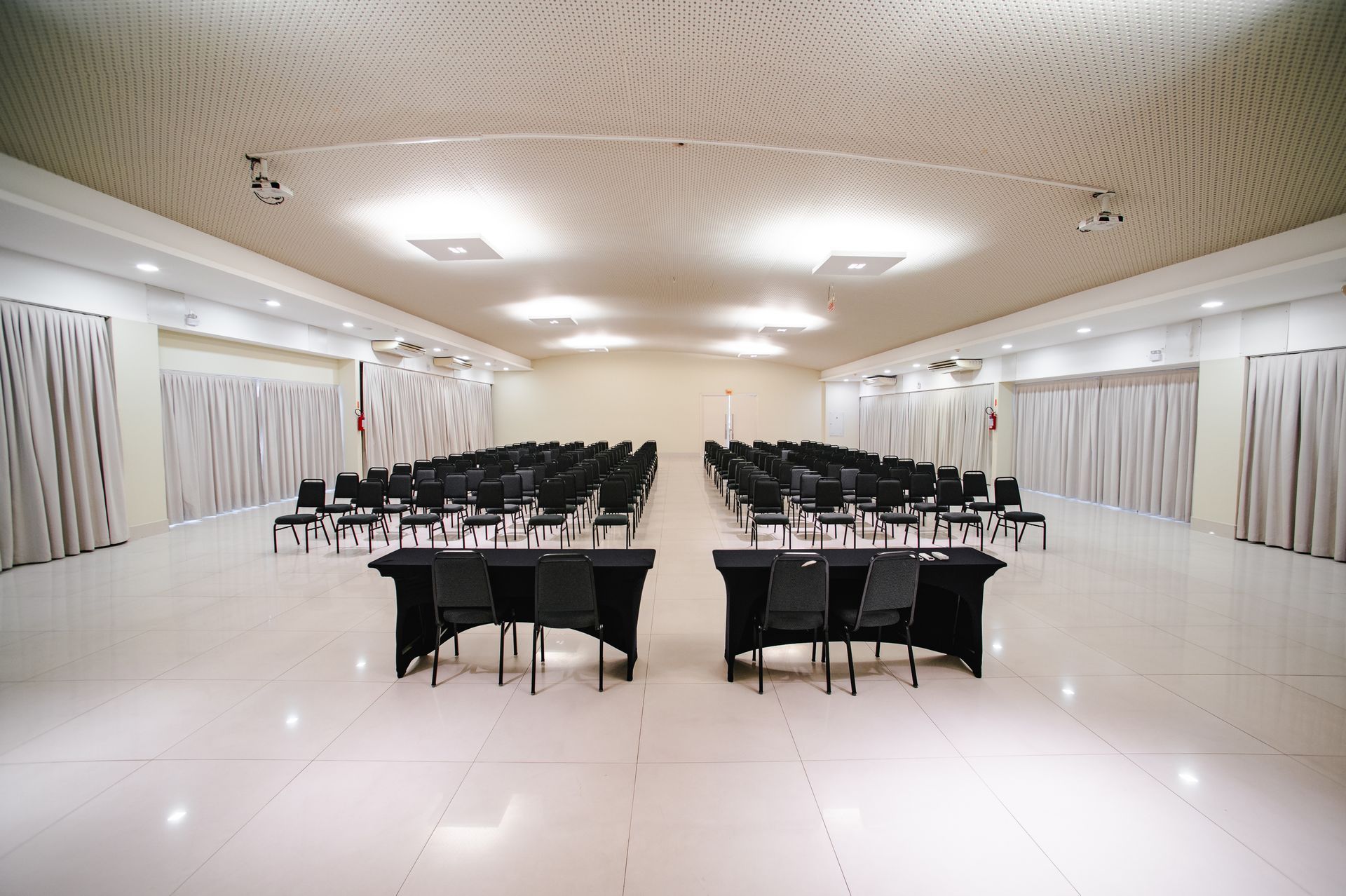 Salão de banquetes vazio com piso de azulejos brancos, luminárias curvas no teto e fileiras de cadeiras e mesas pretas.
