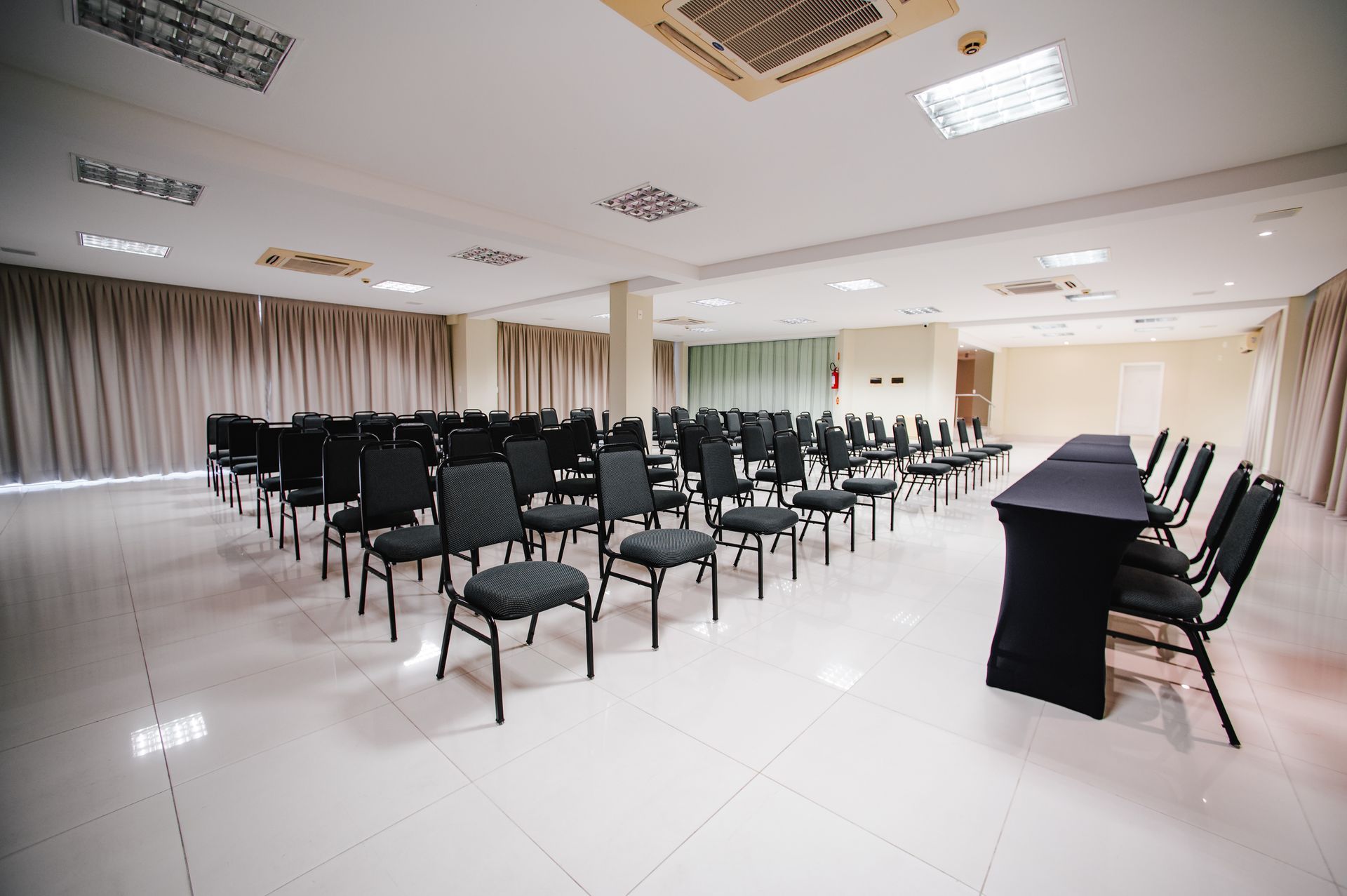 Sala de conferências vazia com fileiras de cadeiras, uma mesa comprida e piso de azulejos brancos sob iluminação forte.