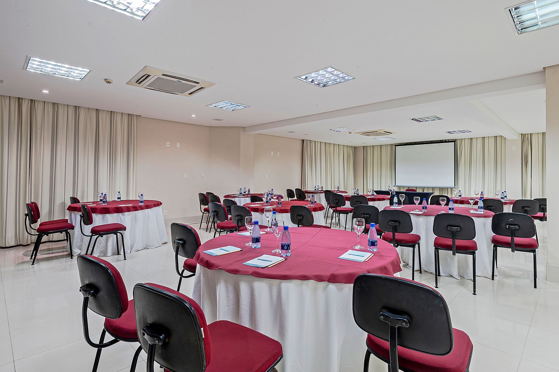Sala de conferências com mesas redondas, cadeiras vermelhas e tela de projeção na frente.