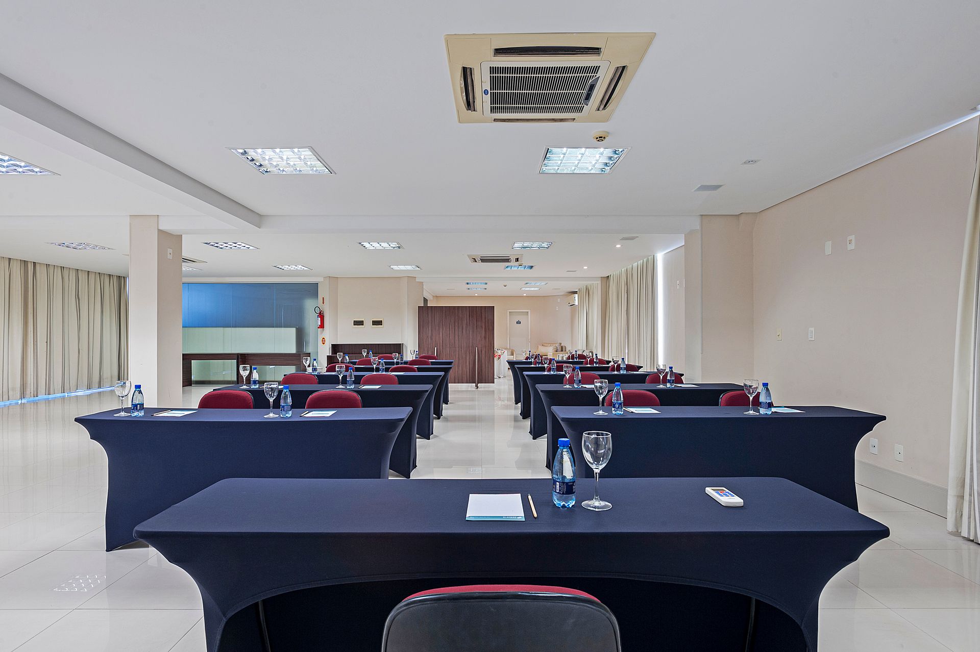 Sala de aula vazia com fileiras de carteiras pretas e cadeiras vermelhas sob luzes fluorescentes brilhantes.