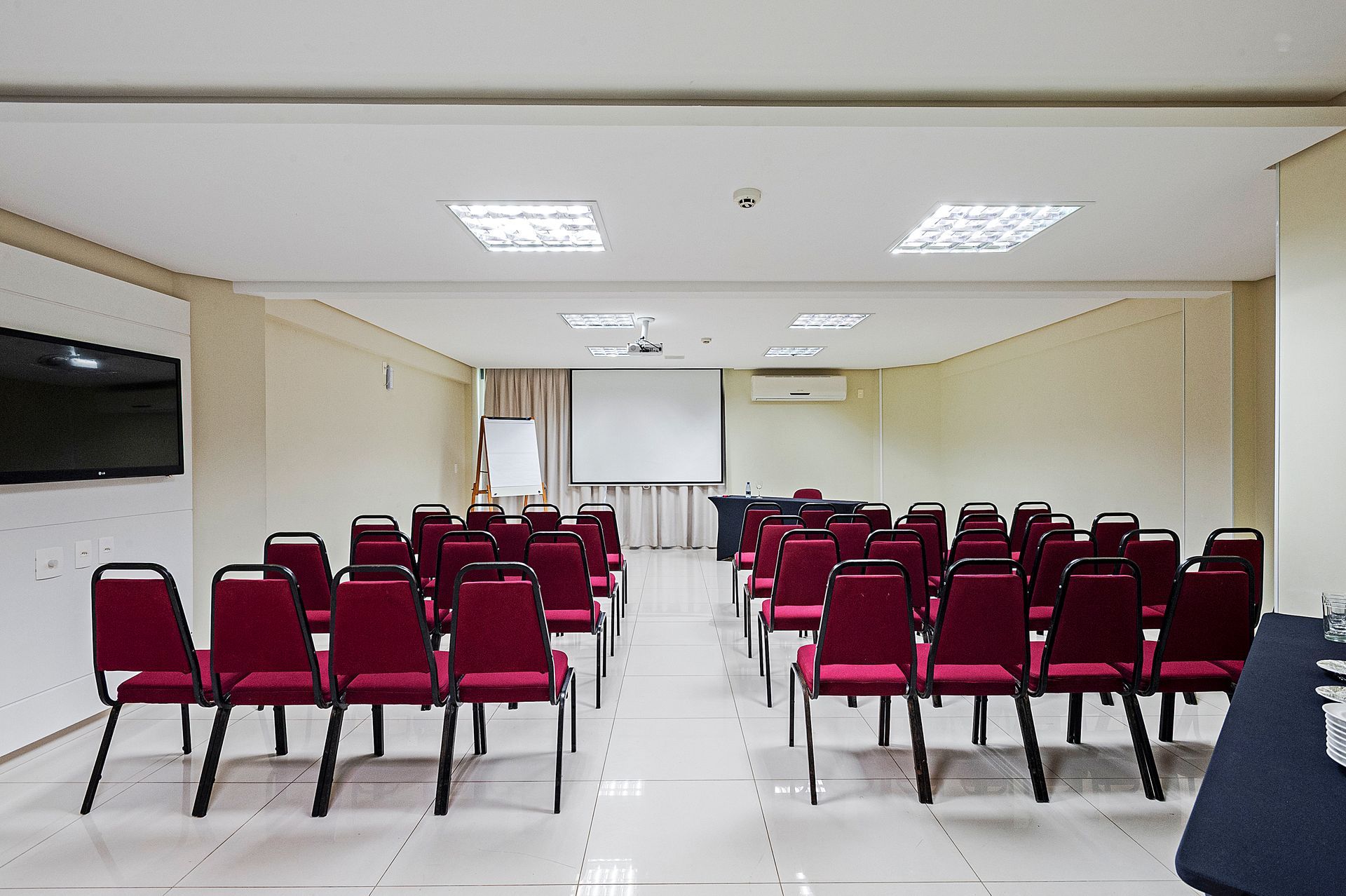 Sala de conferências vazia com fileiras de cadeiras vermelhas voltadas para uma tela de projeção.