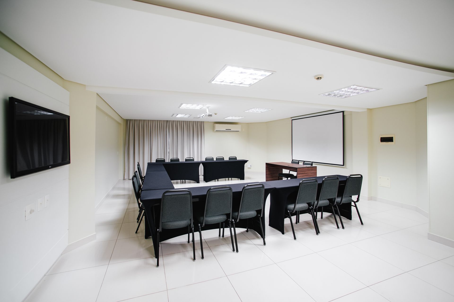 Sala de conferências vazia com cadeiras pretas, mesa branca, TV de parede e tela de projeção.