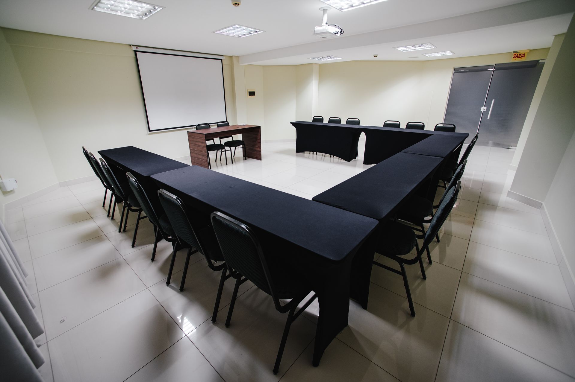 Mesa de conferência preta em formato de U em uma sala de reuniões vazia, com tela de projeção e cadeiras.