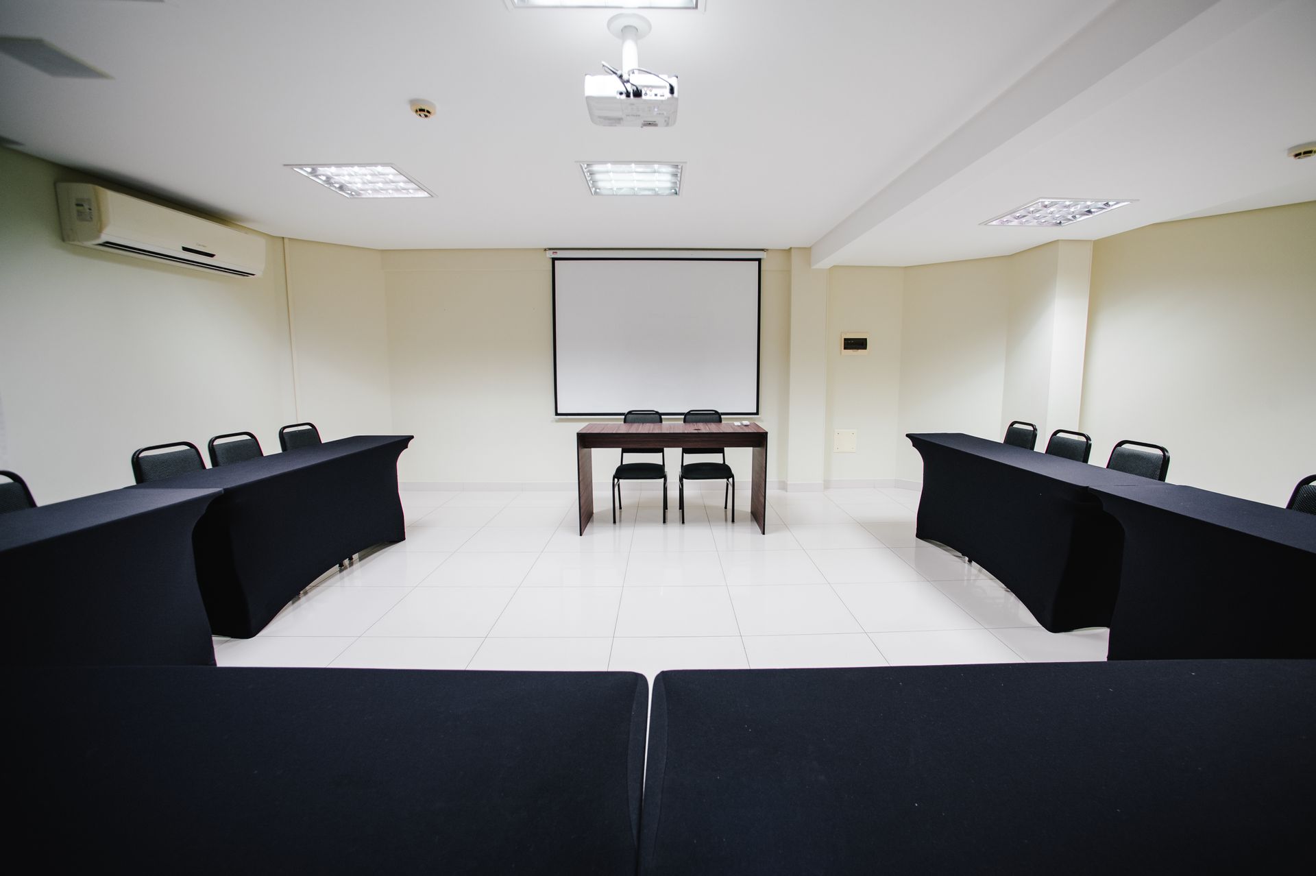 Sala de conferências vazia com mesas pretas em formato de U e uma tela de projeção na frente.
