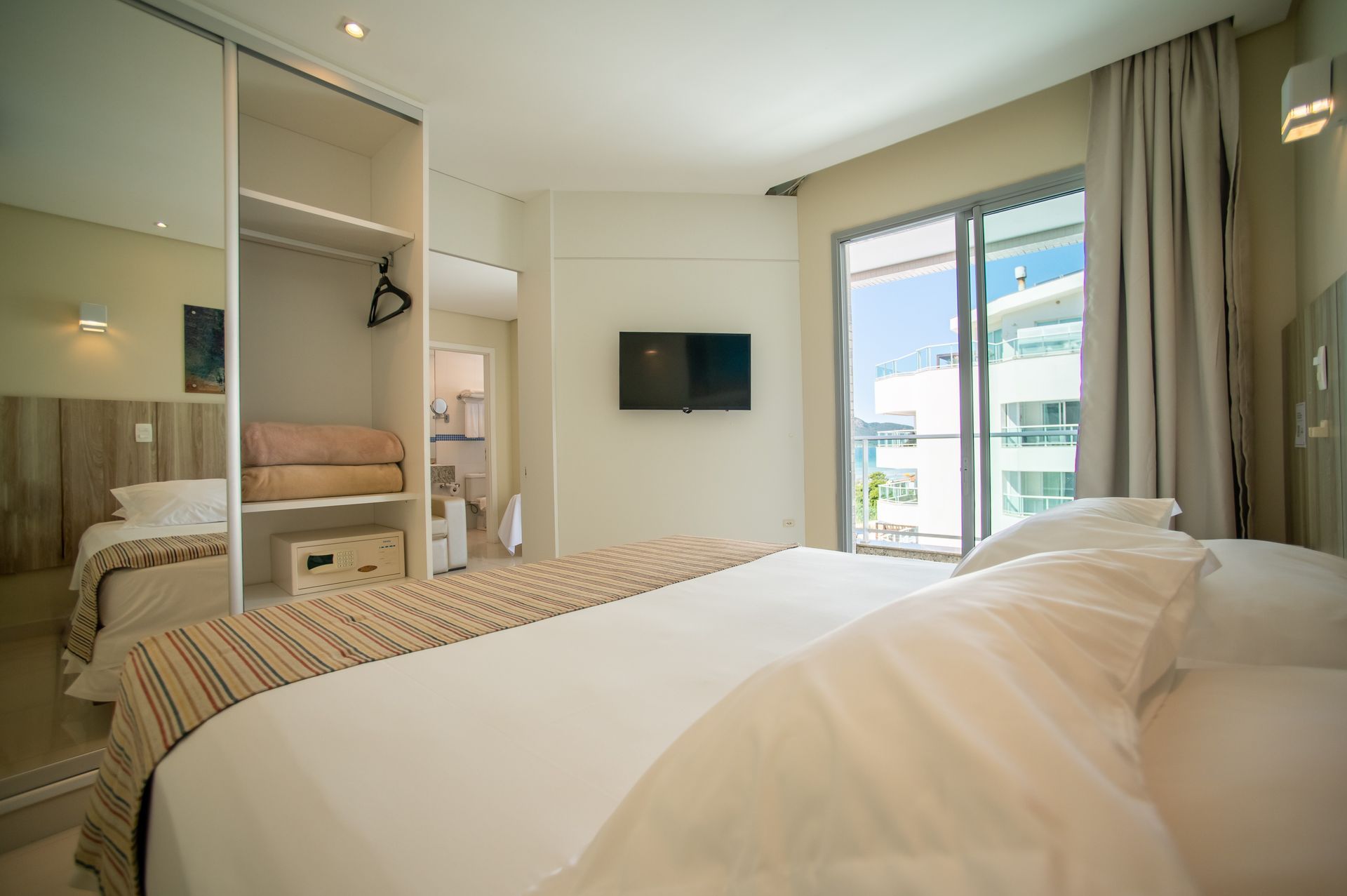 Quarto moderno com cama, armário embutido, TV de parede e portas de varanda com cortinas.