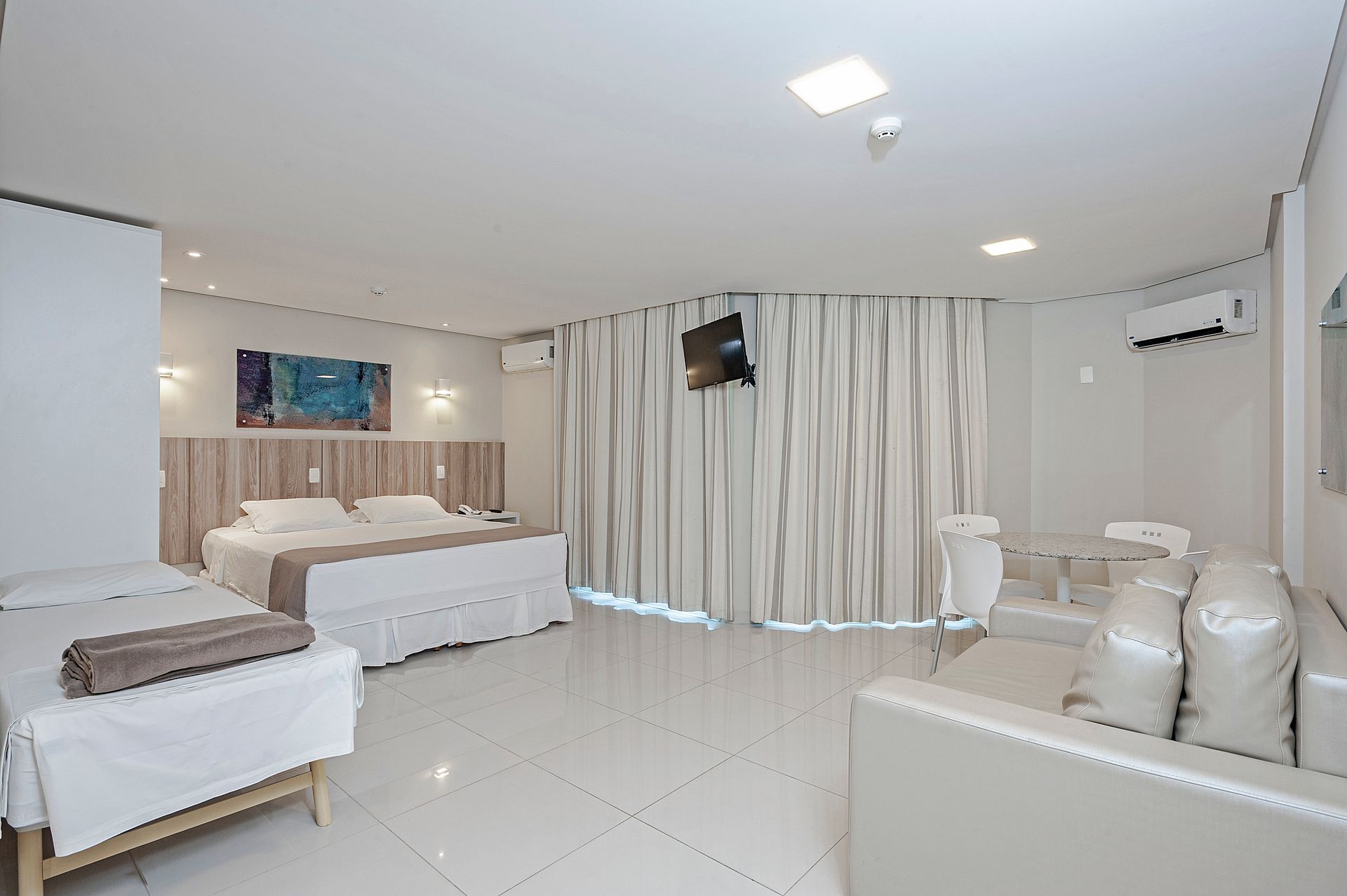 Quarto de hotel branco e luminoso com duas camas, cortinas, piso brilhante e área de estar.