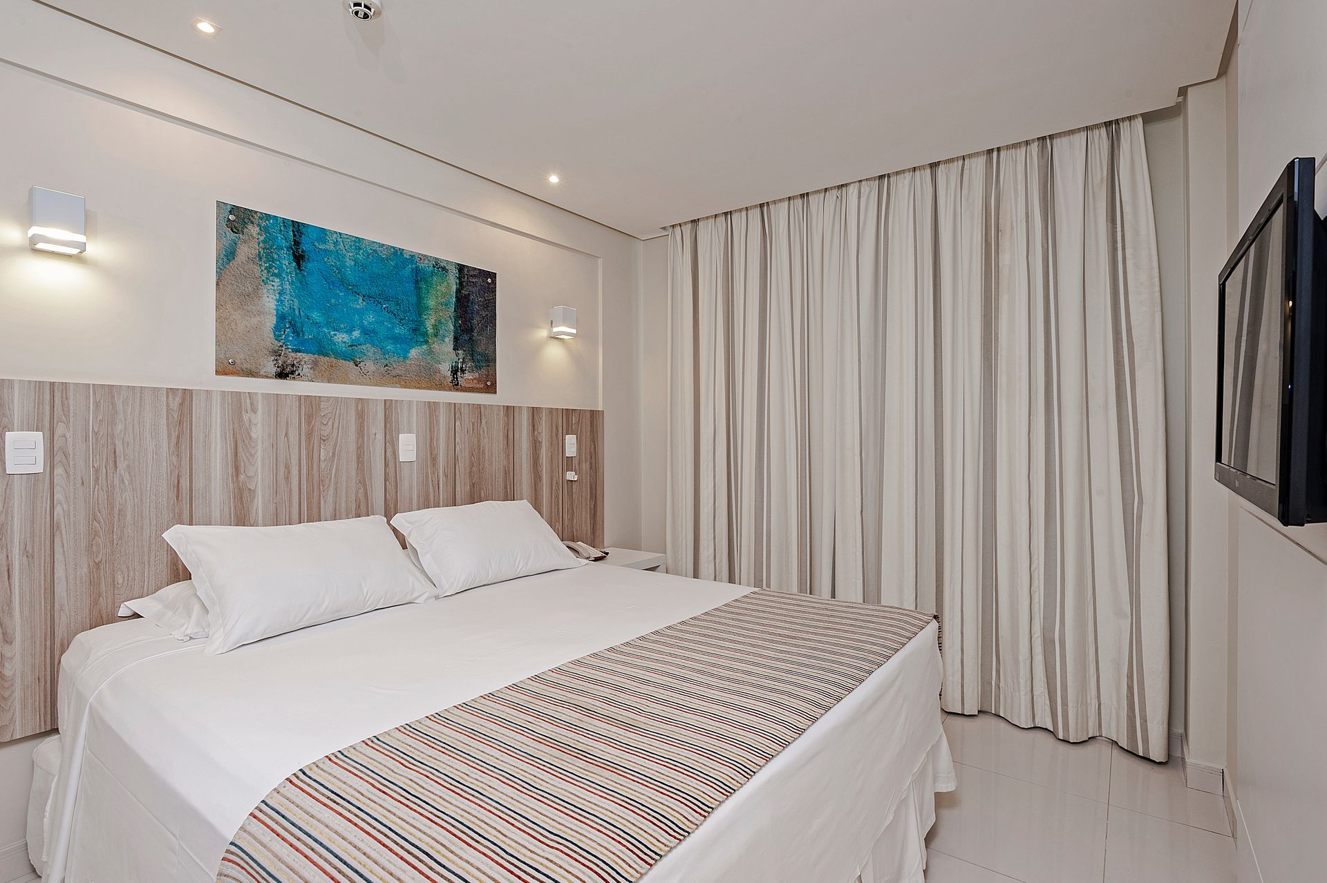Quarto moderno com cama branca, cabeceira de madeira, arte azul na parede e cortinas do chão ao teto.