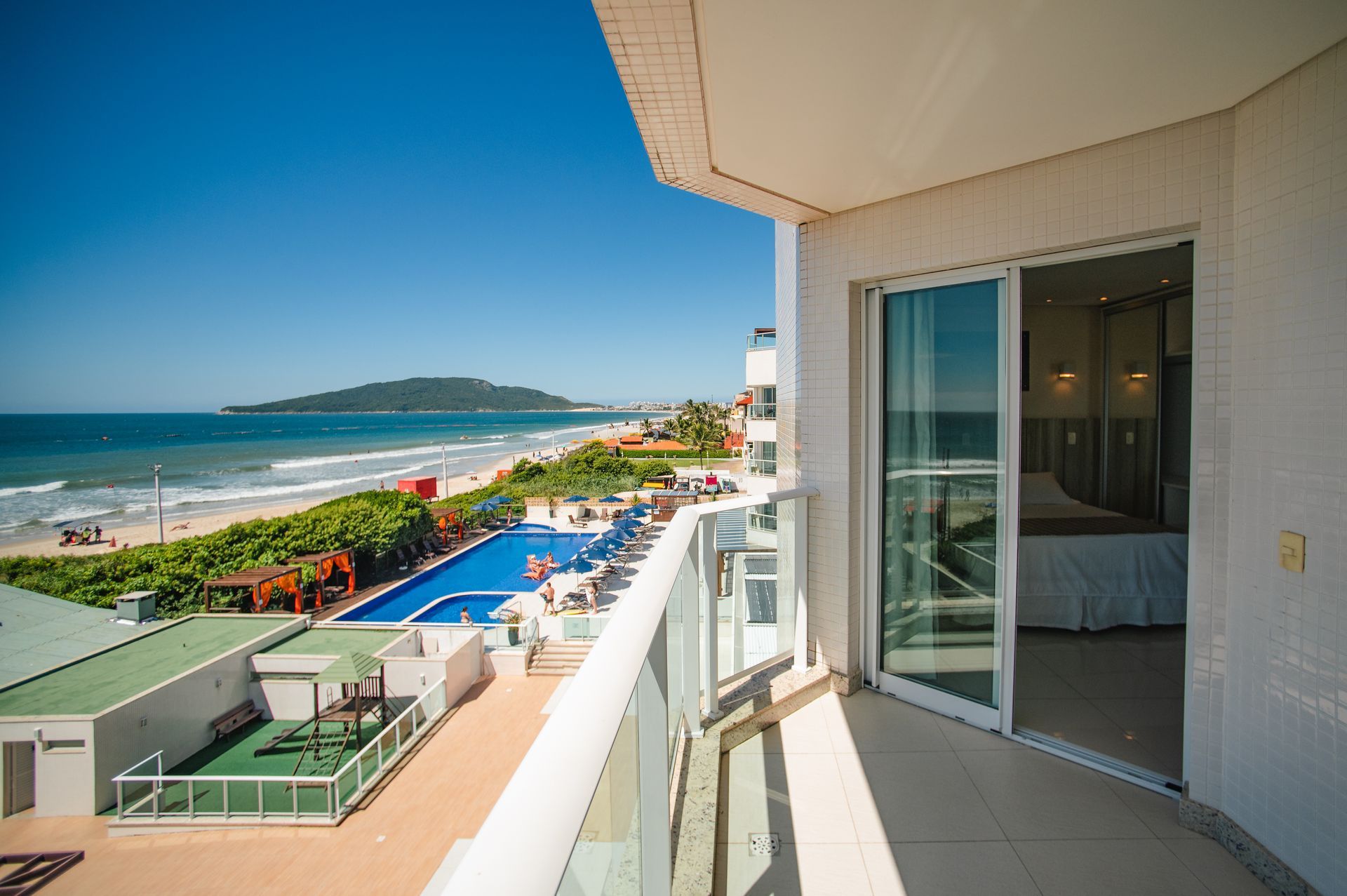 Vista da varanda de um resort de praia com oceano, piscina e litoral ao longe sob um céu azul claro.