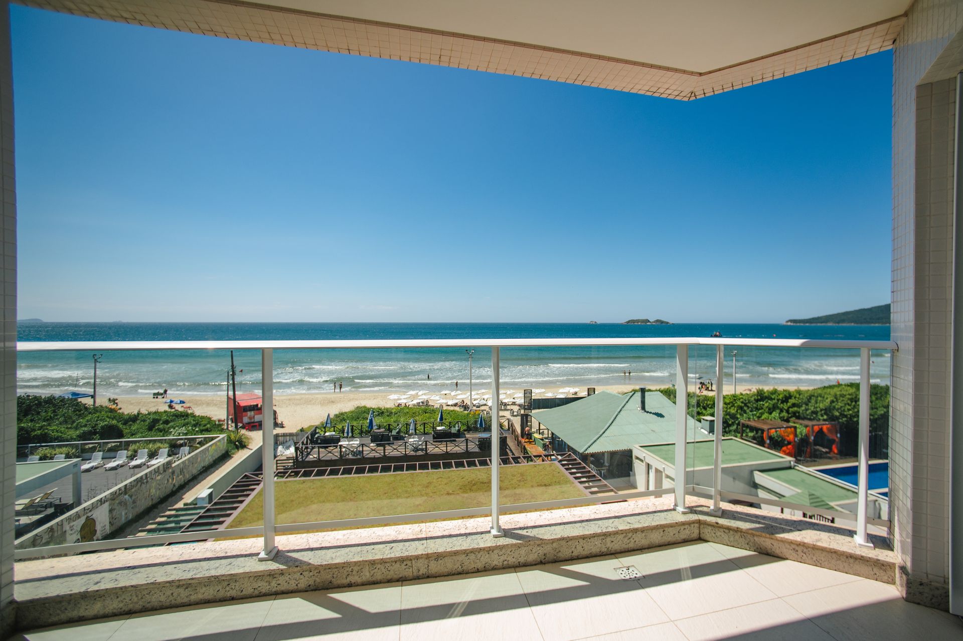 Vista da varanda para um oceano azul brilhante, praia de areia e litoral distante sob um céu limpo.