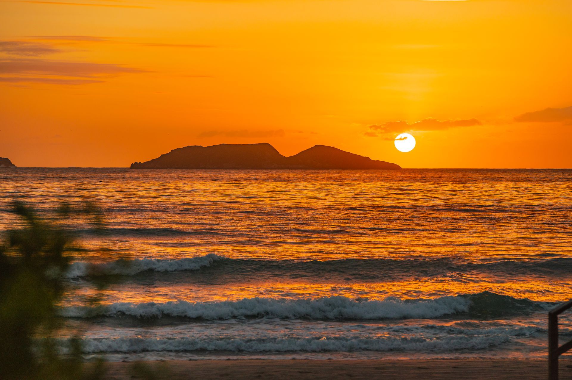 Pôr do sol alaranjado sobre o oceano, com ondas e a silhueta de uma ilha ao longe.
