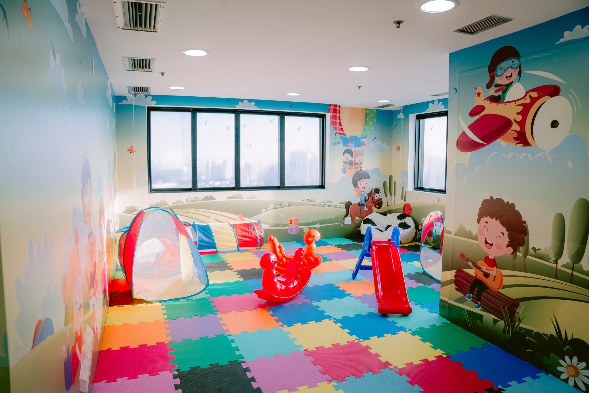 Sala de jogos infantil colorida com tapetes de espuma, brinquedos, murais nas paredes e janelas amplas.