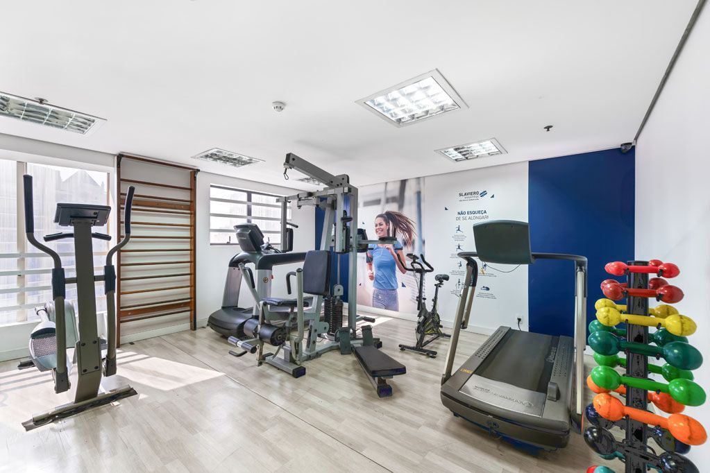 Sala de ginástica iluminada com aparelhos de ginástica, esteira, pesos e uma parede de destaque azul.