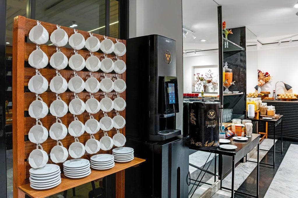 Interior de café com pratos brancos empilhados, prateleiras pretas e cadeiras laranjas sobre um piso de azulejos.