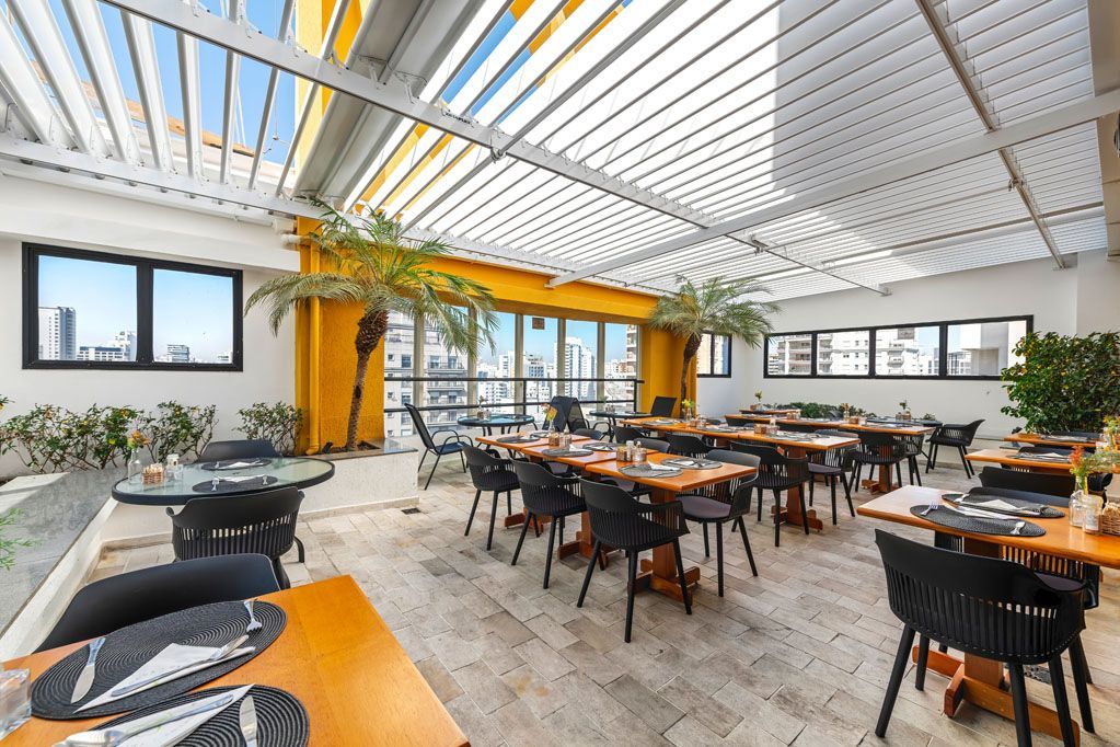 Restaurante luminoso e moderno com mesas de madeira, cadeiras pretas, plantas e teto com claraboia.