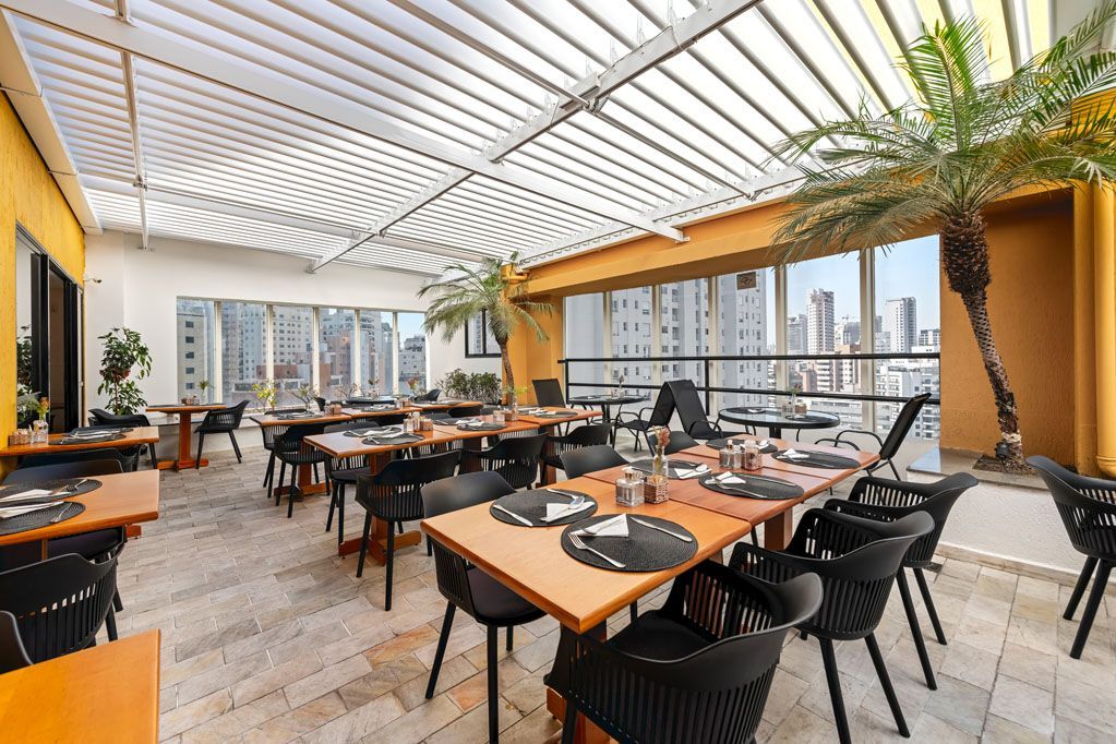 Restaurante na cobertura com cadeiras pretas, mesas de madeira e vista para o horizonte da cidade sob um toldo ripado.