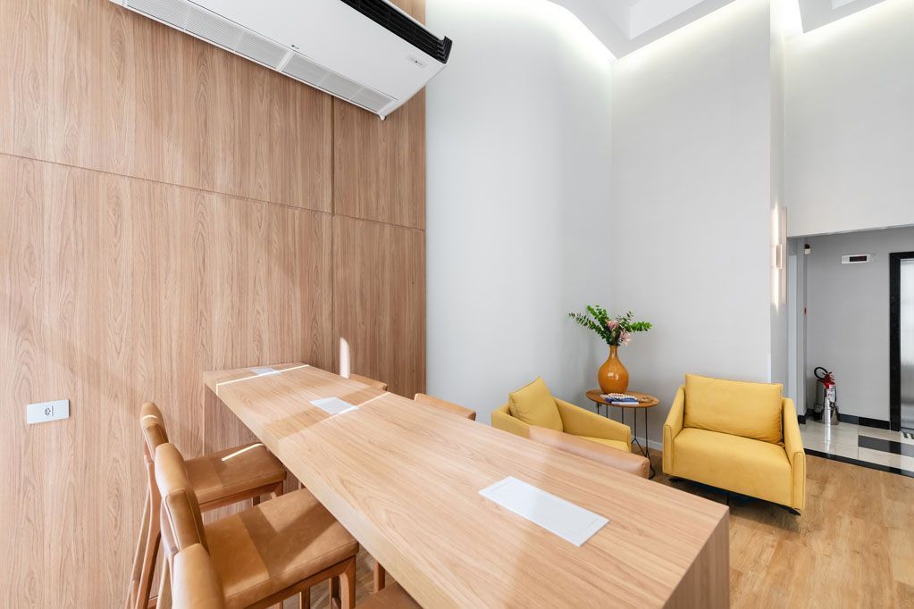 Sala de reuniões moderna com mesa comprida de madeira, cadeiras bege, poltronas amarelas e um vaso de plantas.