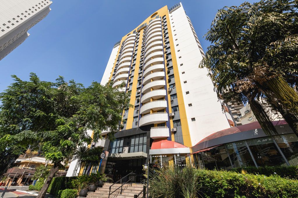 Prédio alto e moderno com apartamentos, detalhes em amarelo e árvores na frente, sob um céu azul.