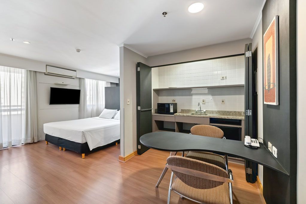 Apartamento estúdio moderno com cama, TV de parede, copa e pequena área para refeições.