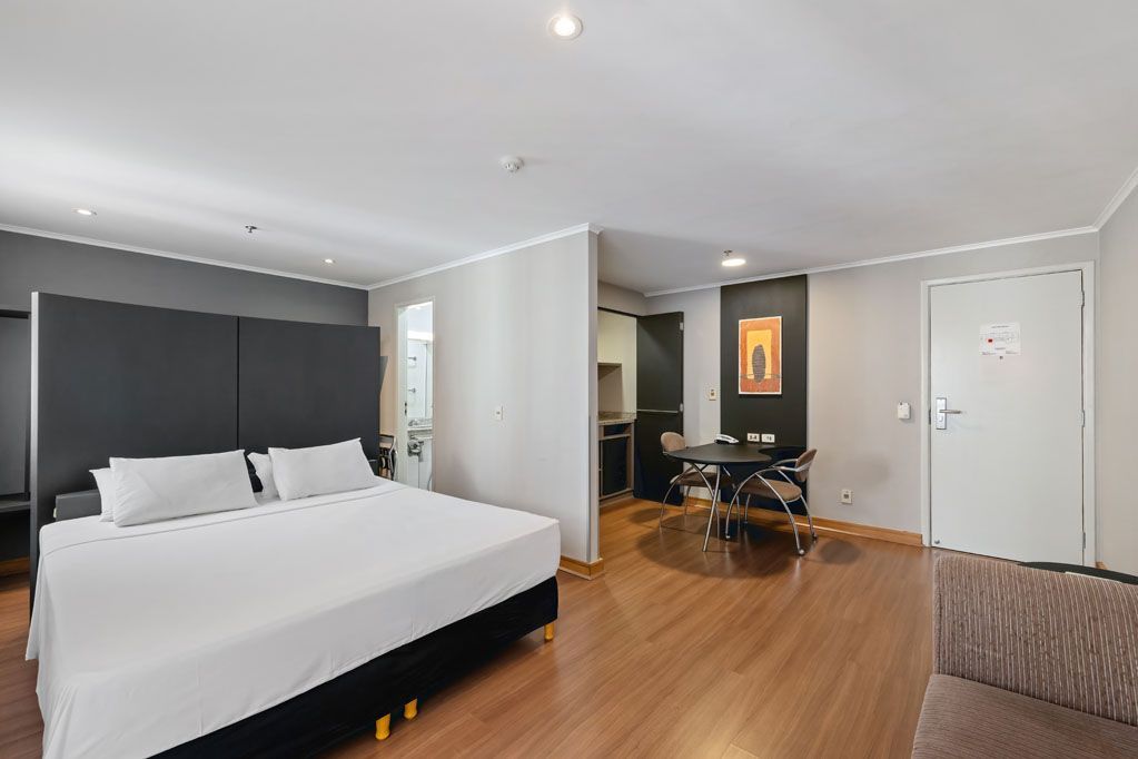 Quarto de hotel moderno com cama grande, piso de madeira, cadeira de escritório e guarda-roupa em tons neutros.