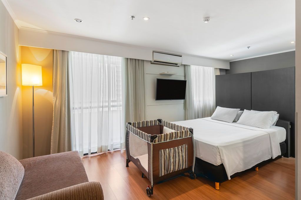 Quarto de hotel moderno com cama, berço, poltrona, TV de parede e janelas amplas com cortinas.