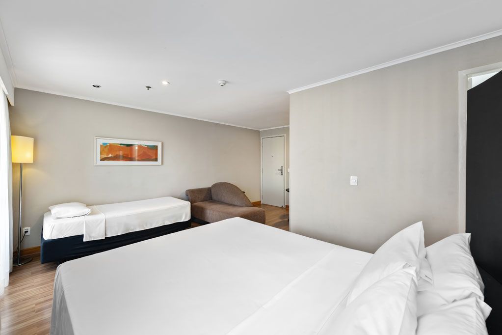 Quarto de hotel luminoso com duas camas, paredes bege, piso de madeira e uma pequena obra de arte laranja na parede.