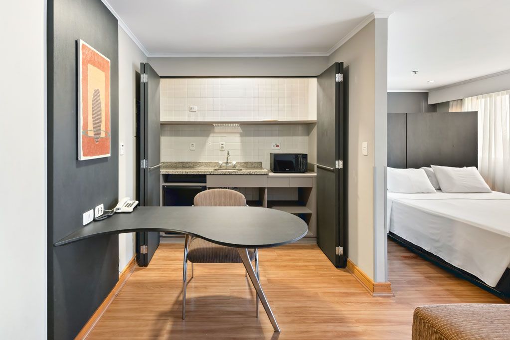 Apartamento estúdio moderno com escrivaninha, cozinha compacta e cama, com piso de madeira e paredes em tons escuros.
