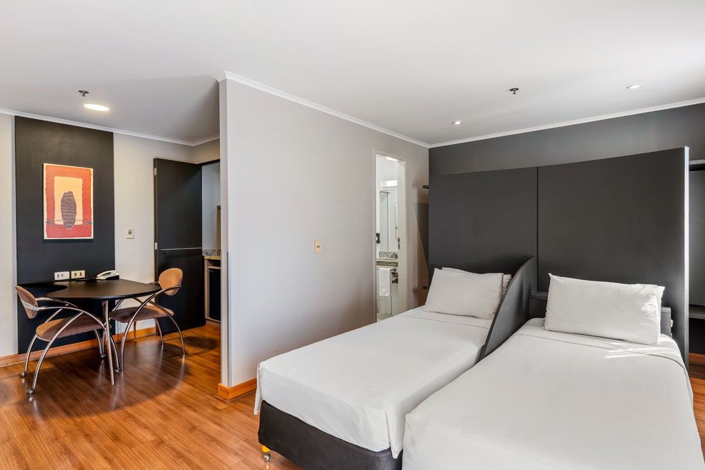Quarto de hotel moderno com duas camas de solteiro, piso de madeira e área de jantar ao fundo.